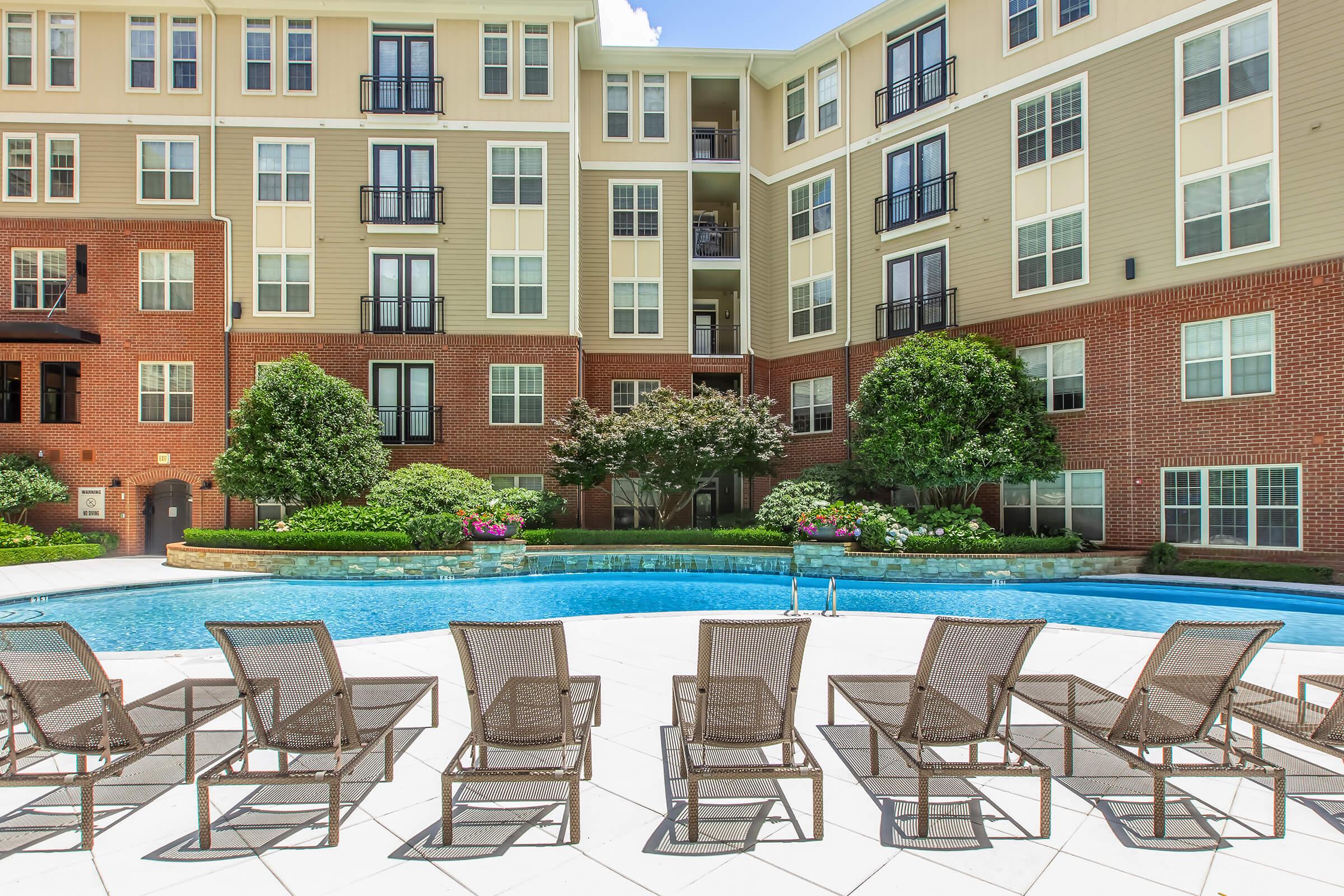 A serene outdoor pool area surrounded by lounge chairs, with landscaped greenery and an apartment building in the background. The pool reflects blue skies and is bordered by colorful flowerbeds, creating a relaxing atmosphere for residents and visitors.