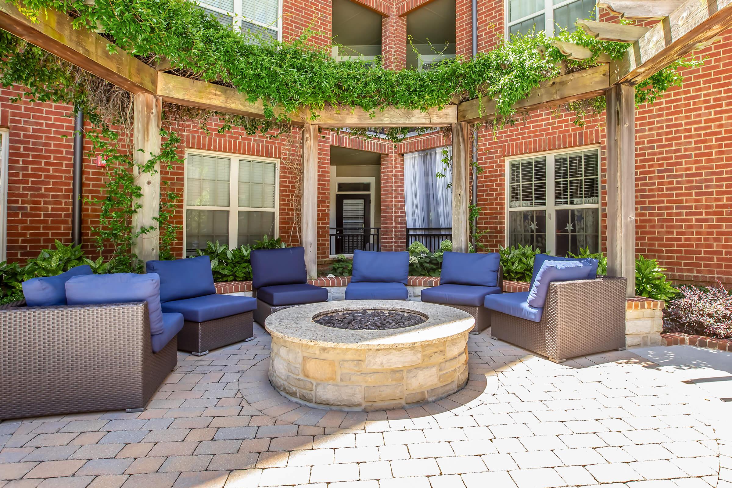 A cozy outdoor seating area featuring blue cushioned chairs arranged around a circular stone fire pit, surrounded by greenery and red brick buildings, offering a welcoming atmosphere for relaxation and social gatherings.