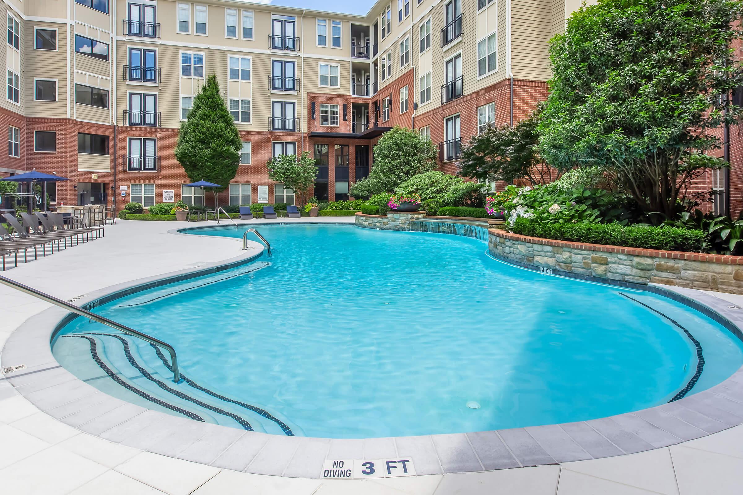 A swimming pool with a curved design in the foreground, surrounded by lounge chairs and greenery. In the background, there are several multi-story apartment buildings with balconies. The pool area features a stone border and clear blue water, with nearby landscaping including shrubs and trees.