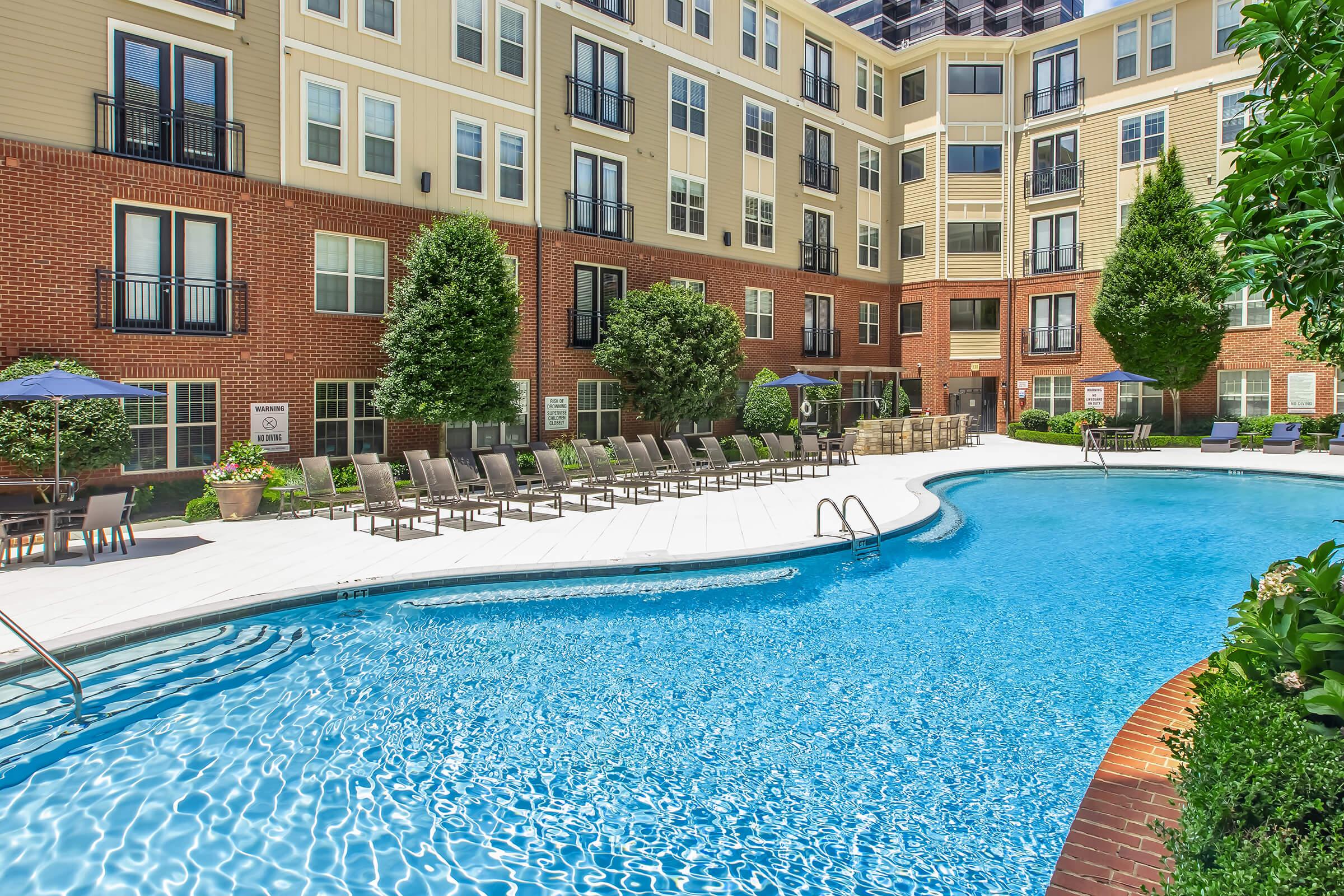 A clear blue swimming pool surrounded by lounge chairs and potted plants, located in a residential complex. The pool area features shaded seating under umbrellas, and the backdrop includes a multi-story brick building with large windows and landscaped greenery.