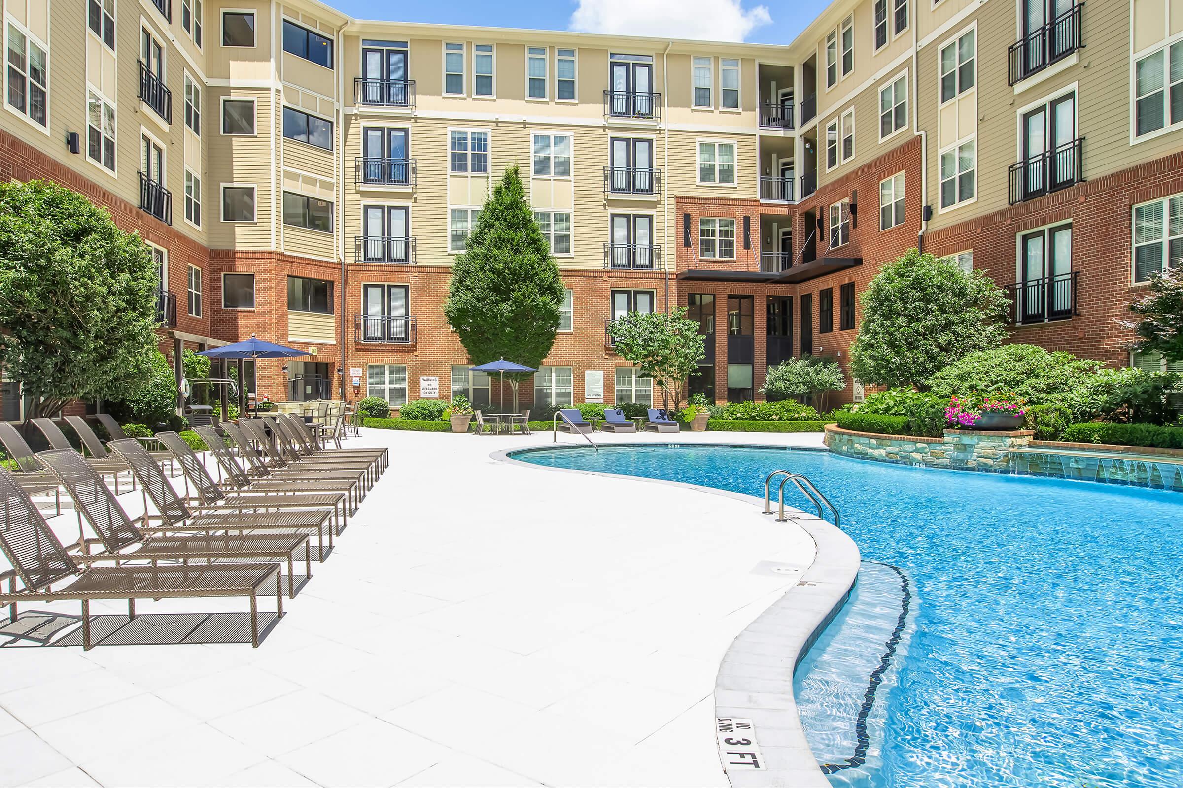 A spacious outdoor pool area surrounded by lounge chairs, with lush landscaping and trees. The pool features a curved edge, and there are shaded seating areas with umbrellas. The backdrop consists of a multi-story apartment building with large windows and balconies under a clear blue sky.