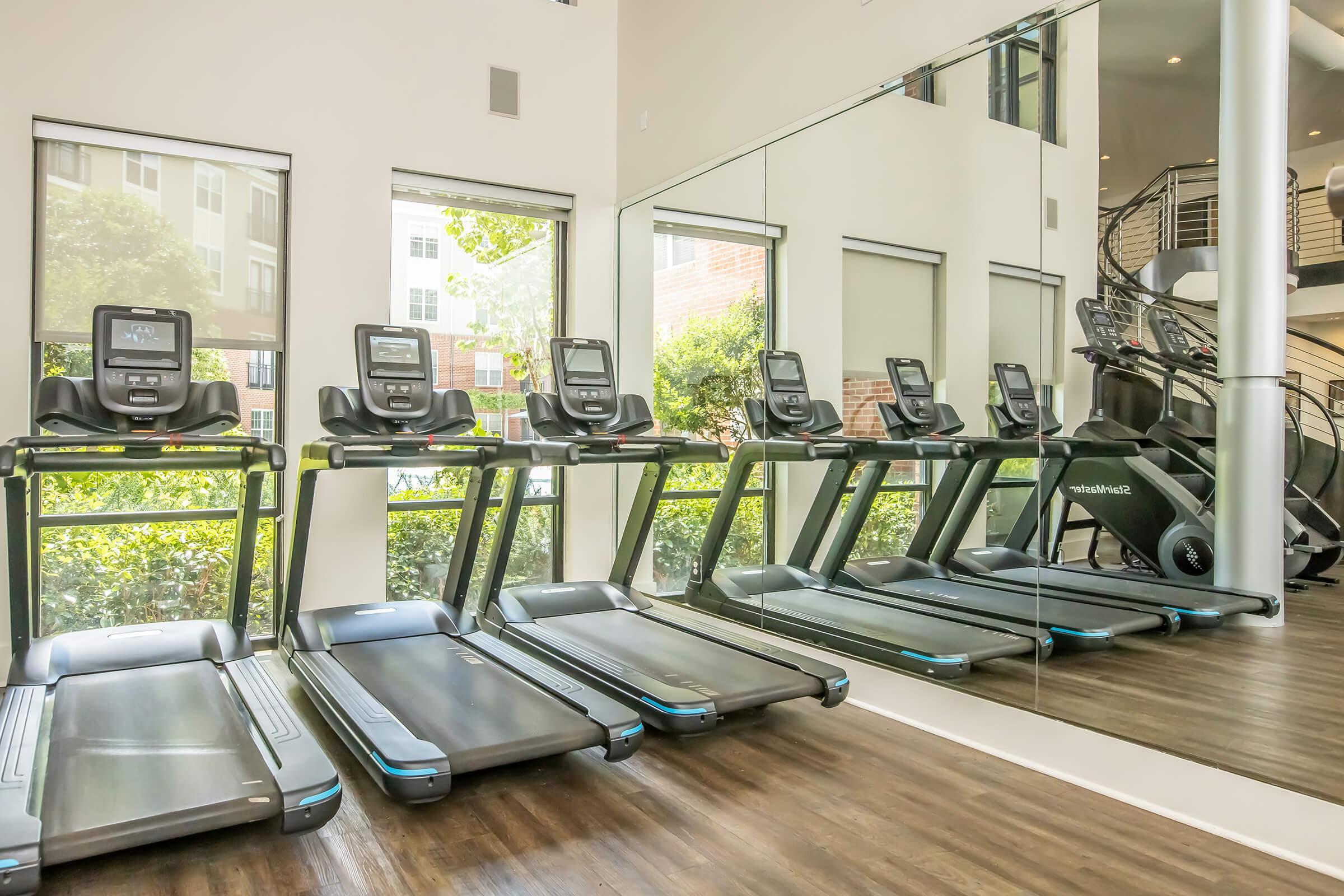 A fitness room featuring several modern treadmills lined up against a mirror. Bright natural light enters through large windows, illuminating the space and showcasing greenery outside. The stylish interior includes hardwood flooring and a glimpse of a staircase in the background.