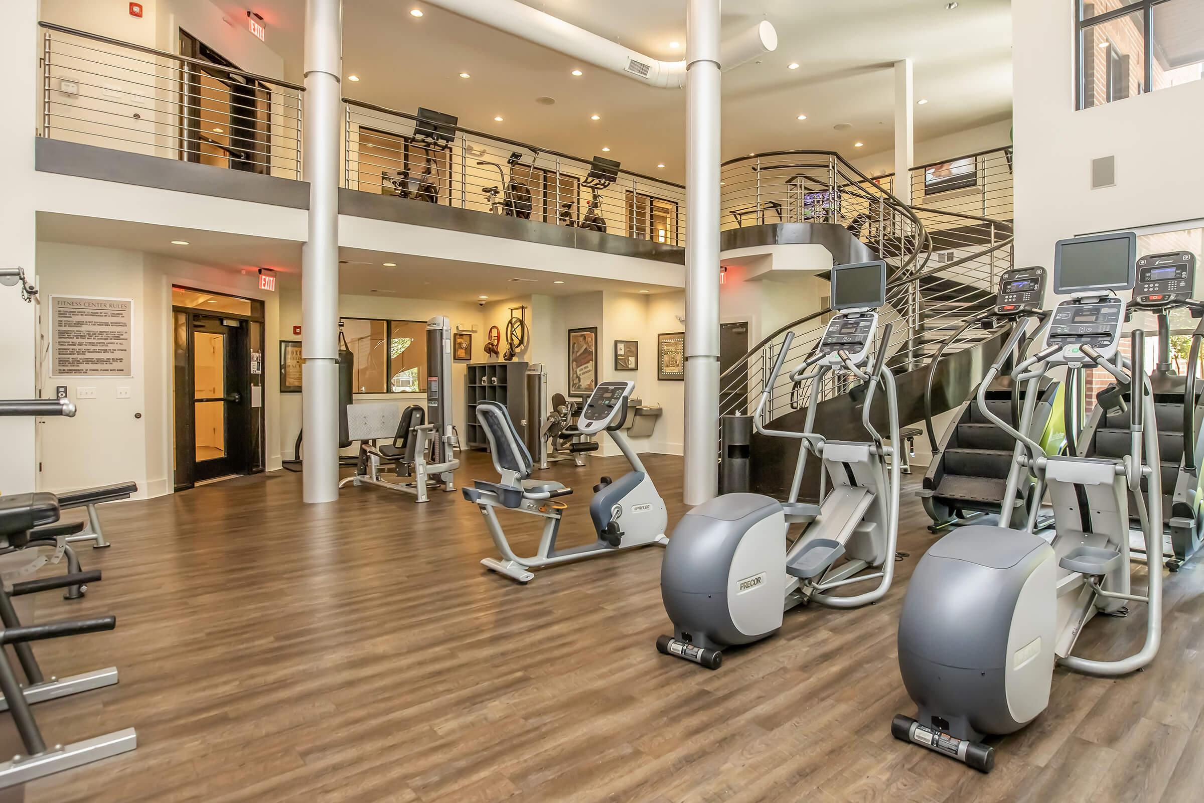 A modern gym interior featuring various exercise equipment, including ellipticals and stationary bikes, arranged on a wooden floor. The space has a high ceiling with natural lighting, a staircase leading to an upper area, and fitness-related decor on the walls.