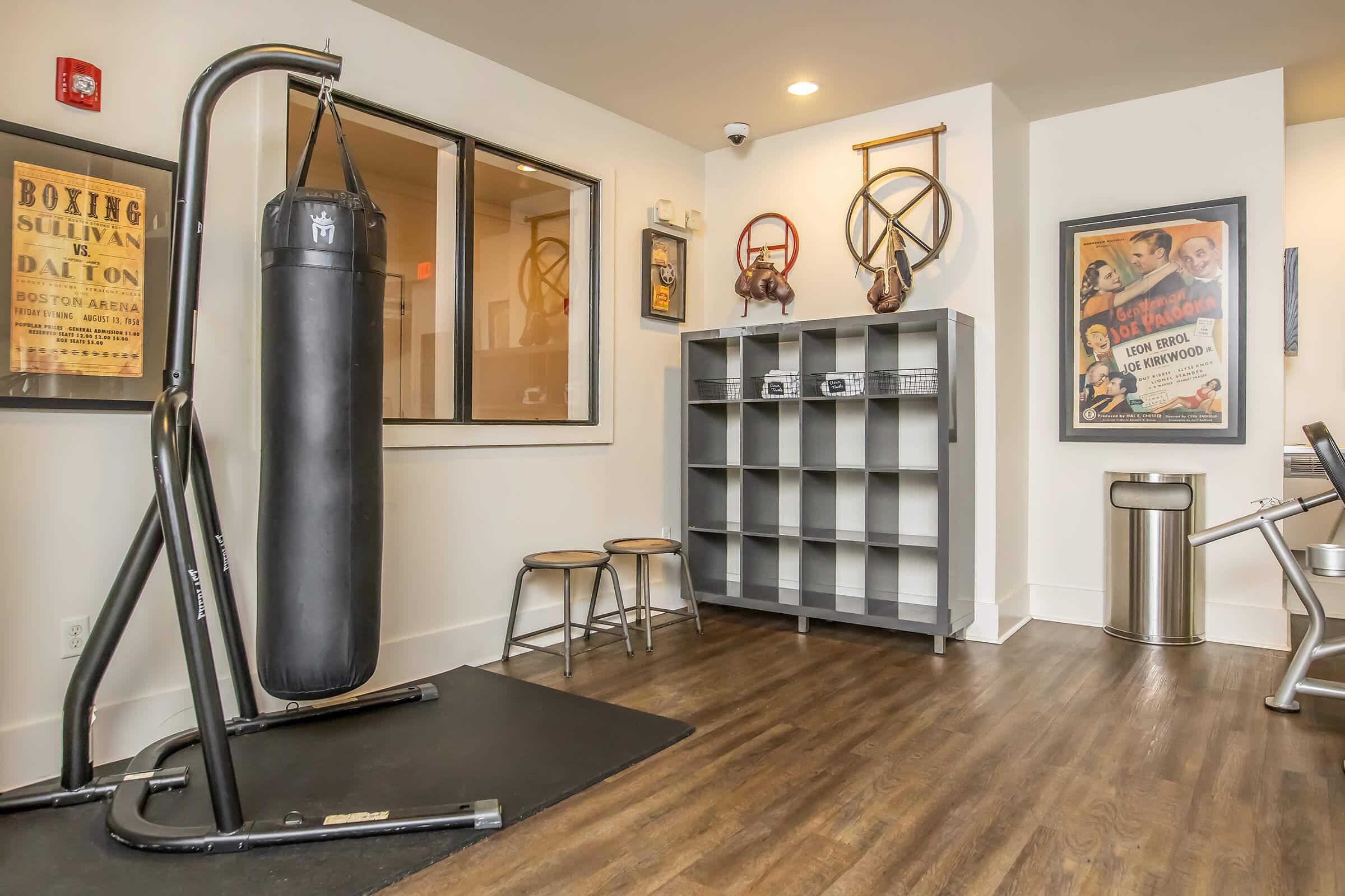 Interior of a fitness room featuring a black punching bag, two metal stools, a gray shelf with fitness equipment, a trash can, and vintage boxing posters on the walls. There's a large window providing natural light.