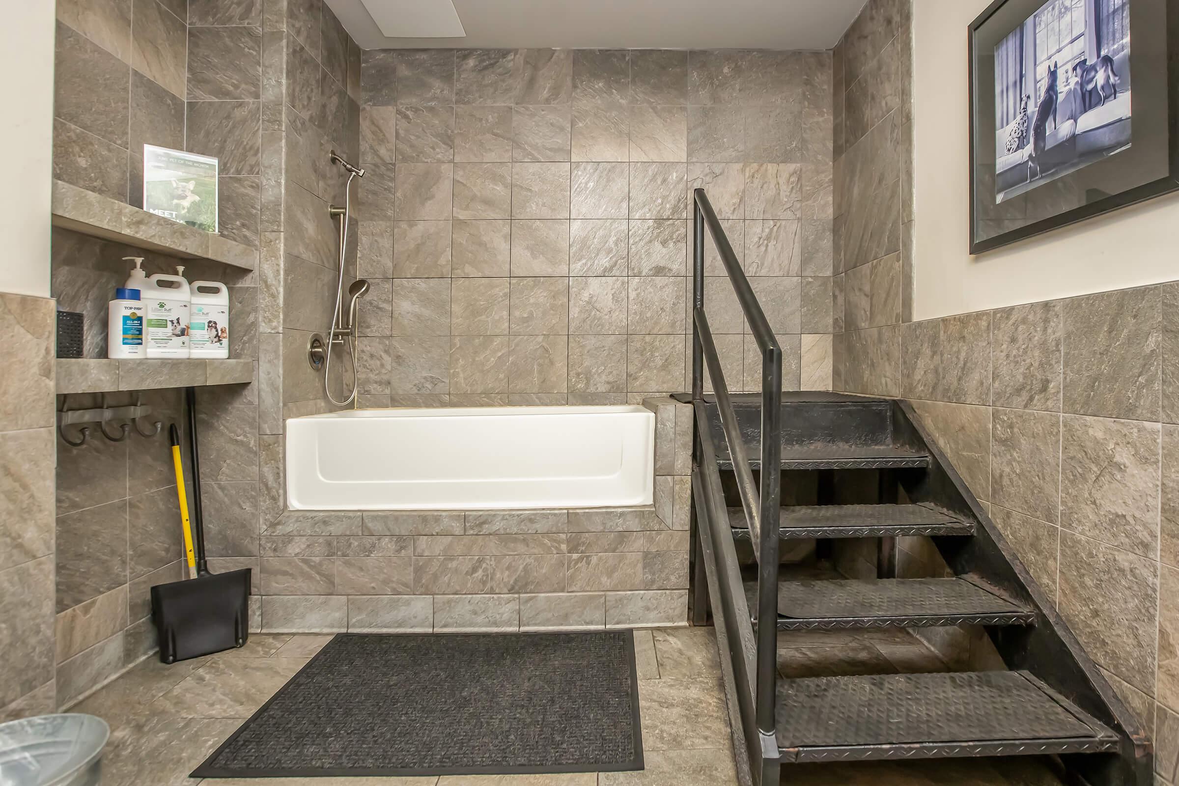 A spacious bathroom featuring a tiled wall, a bathtub with a showerhead, and a set of black stairs. To the side, there are shelves with cleaning products and a few hooks. The floor is covered with a black mat, and there’s a framed picture on the wall.