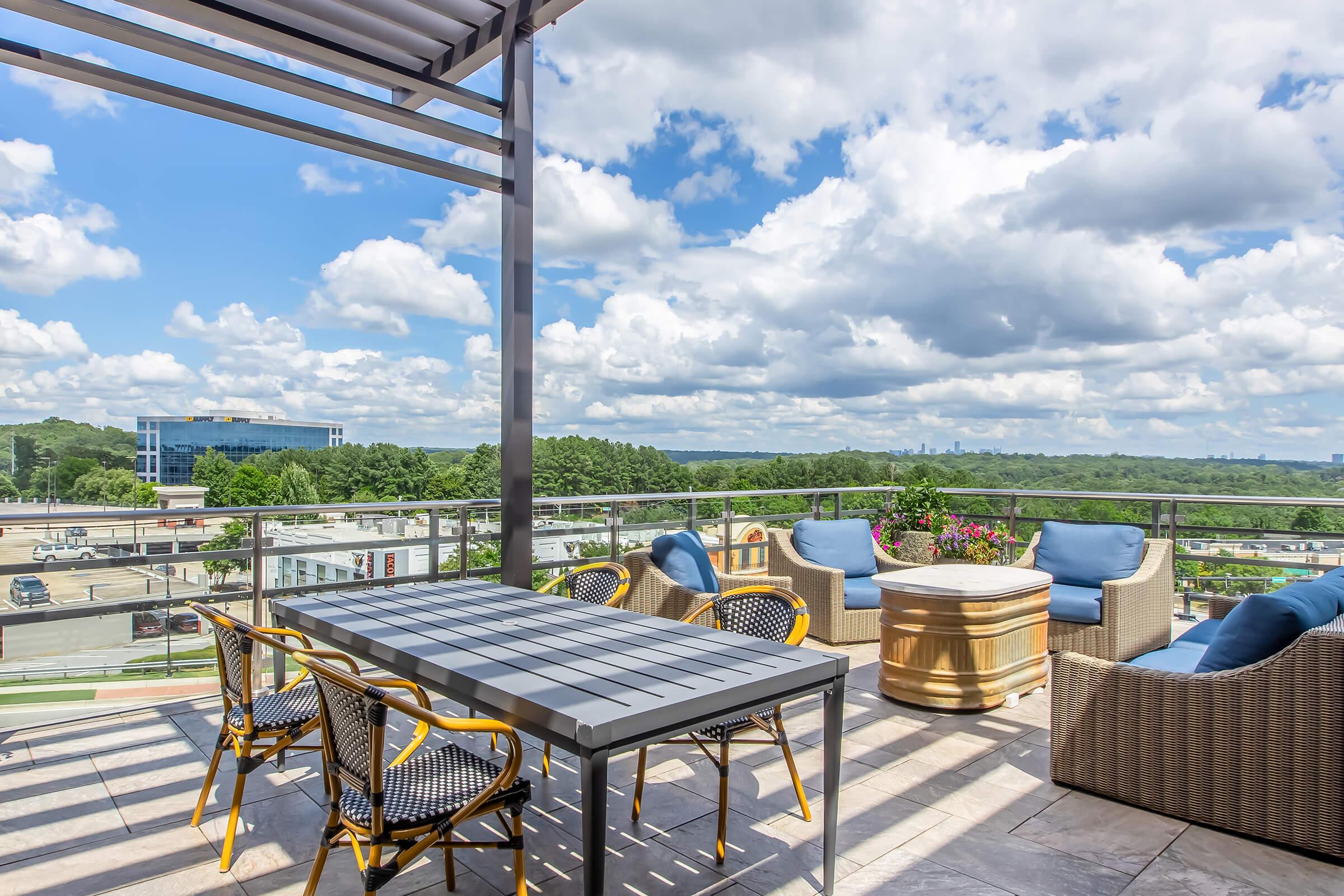 A spacious outdoor terrace featuring a dining table with yellow-accented chairs, comfortable seating areas with cushions, and beautiful views of a blue sky filled with clouds. The background includes greenery and urban elements, creating a relaxing atmosphere.