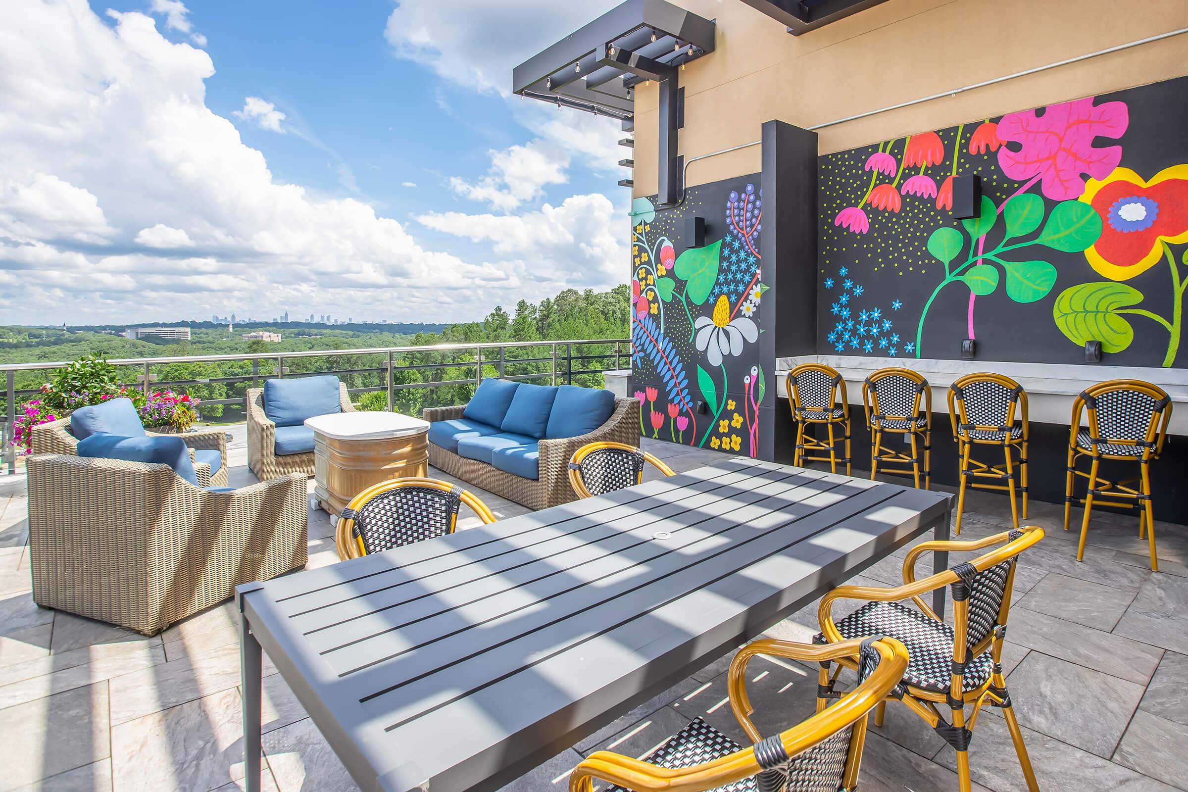 Outdoor rooftop seating area with stylish furniture, including blue chairs and a large dining table. A colorful mural featuring flowers and plants adorns one wall, while lush greenery and a distant skyline are visible in the background under a partly cloudy sky.