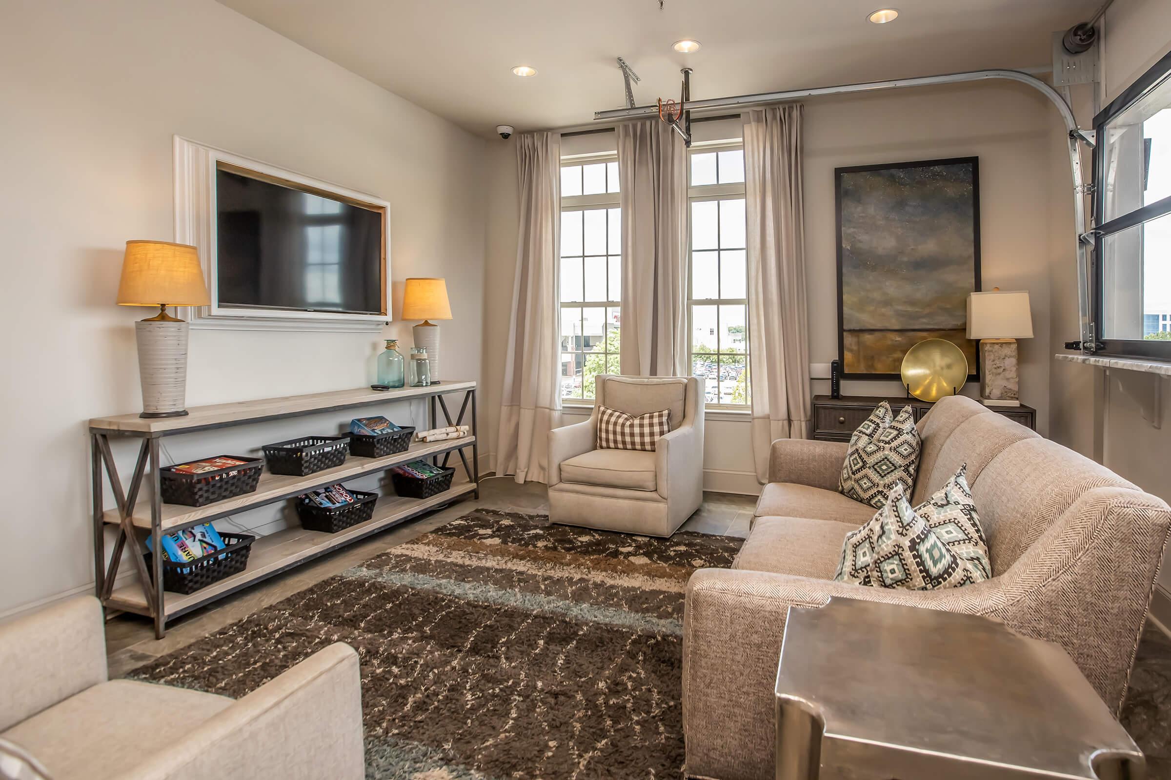 A modern living room with a large window allowing natural light. It features a comfortable beige sofa and armchair, decorative cushions, a coffee table, and an entertainment center with a TV. Soft lighting from table lamps complements the neutral color palette and contemporary art on the wall. A textured area rug adds warmth to the space.