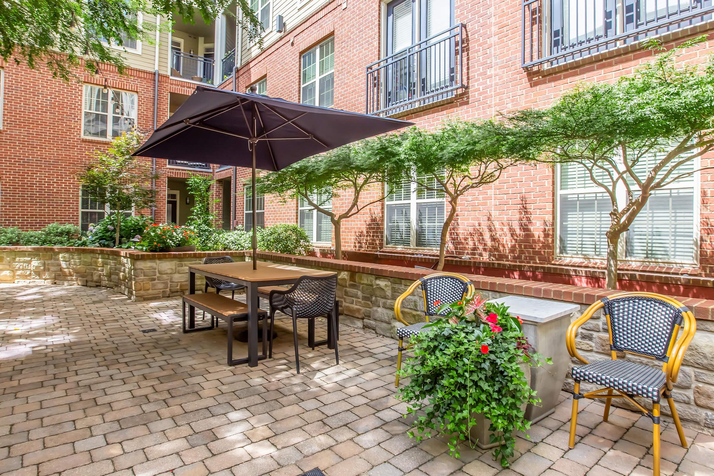 Outdoor patio area featuring a stone-paved floor, a table with an umbrella, and several chairs. Lush greenery and flowering plants surround the seating area, providing a pleasant and inviting atmosphere in a residential setting. Ideal for relaxing or social gatherings.