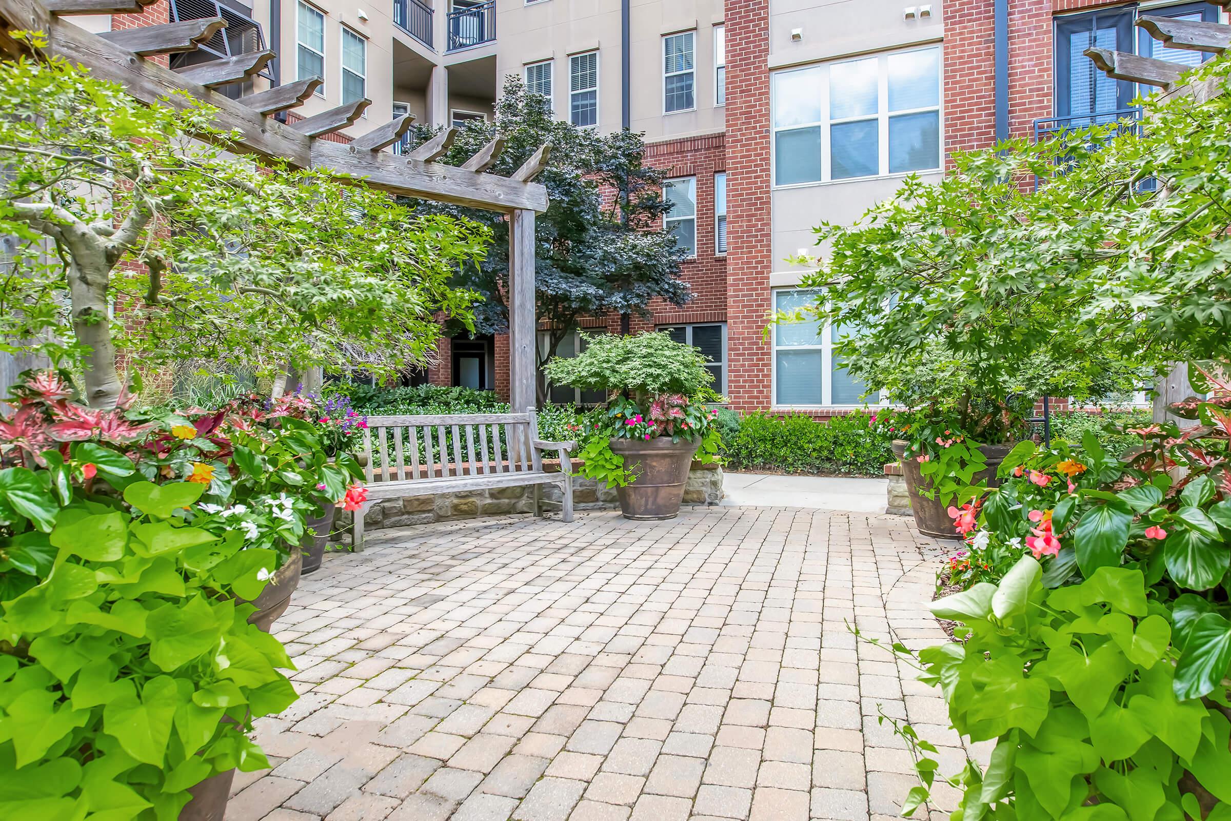 A serene outdoor garden space featuring a wooden pergola, surrounded by lush greenery and colorful flowering plants. A stone pathway leads to a wooden bench, creating a tranquil spot for relaxation. Modern apartment buildings provide a backdrop to this picturesque scene, enhancing the inviting atmosphere.