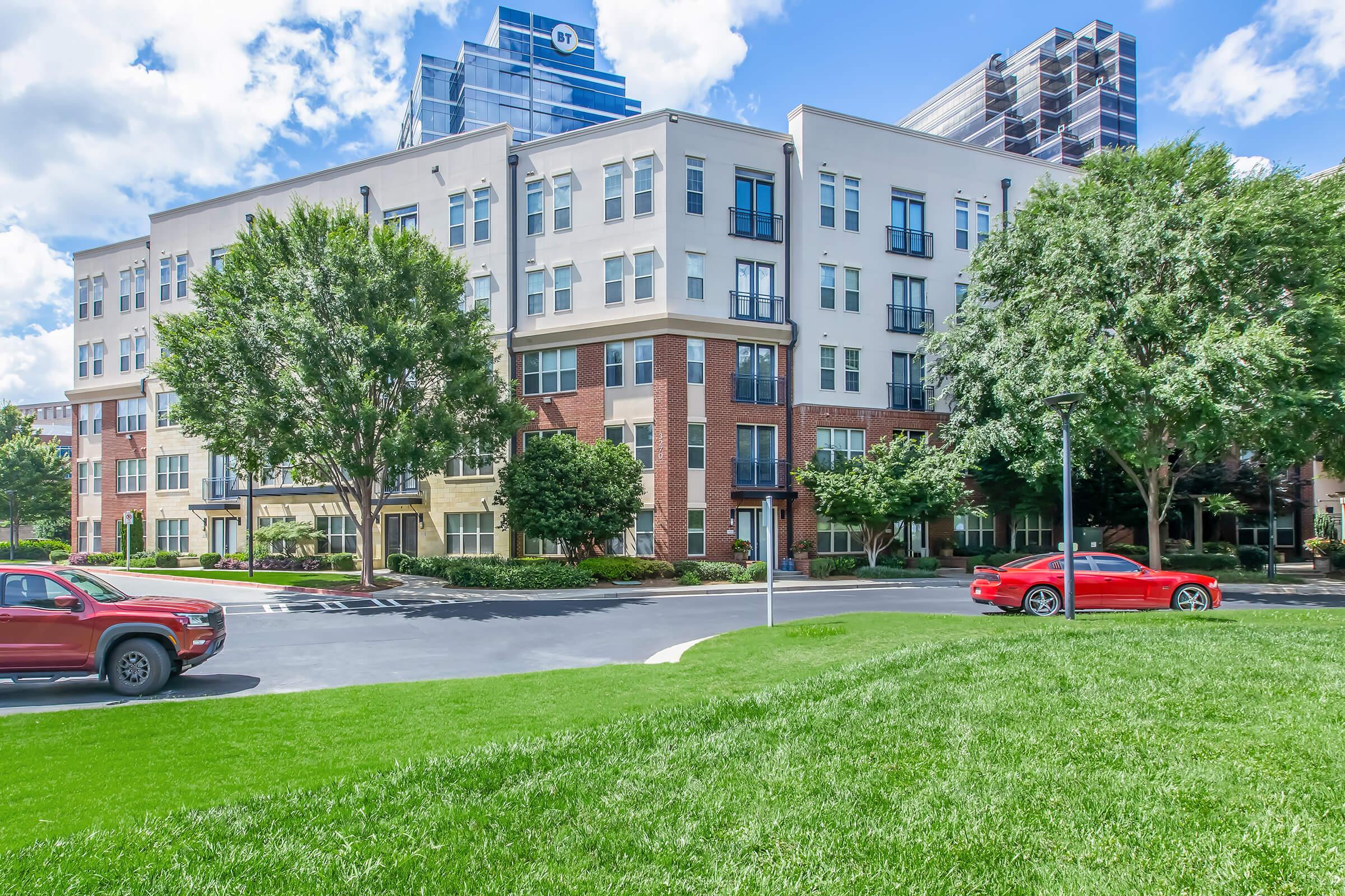A modern apartment building featuring a mix of brick and light-colored facades, surrounded by lush green lawns and trees. A red car is parked in the foreground, and several cars are visible in the parking area. Bright blue sky with fluffy white clouds adds to the vibrant atmosphere.