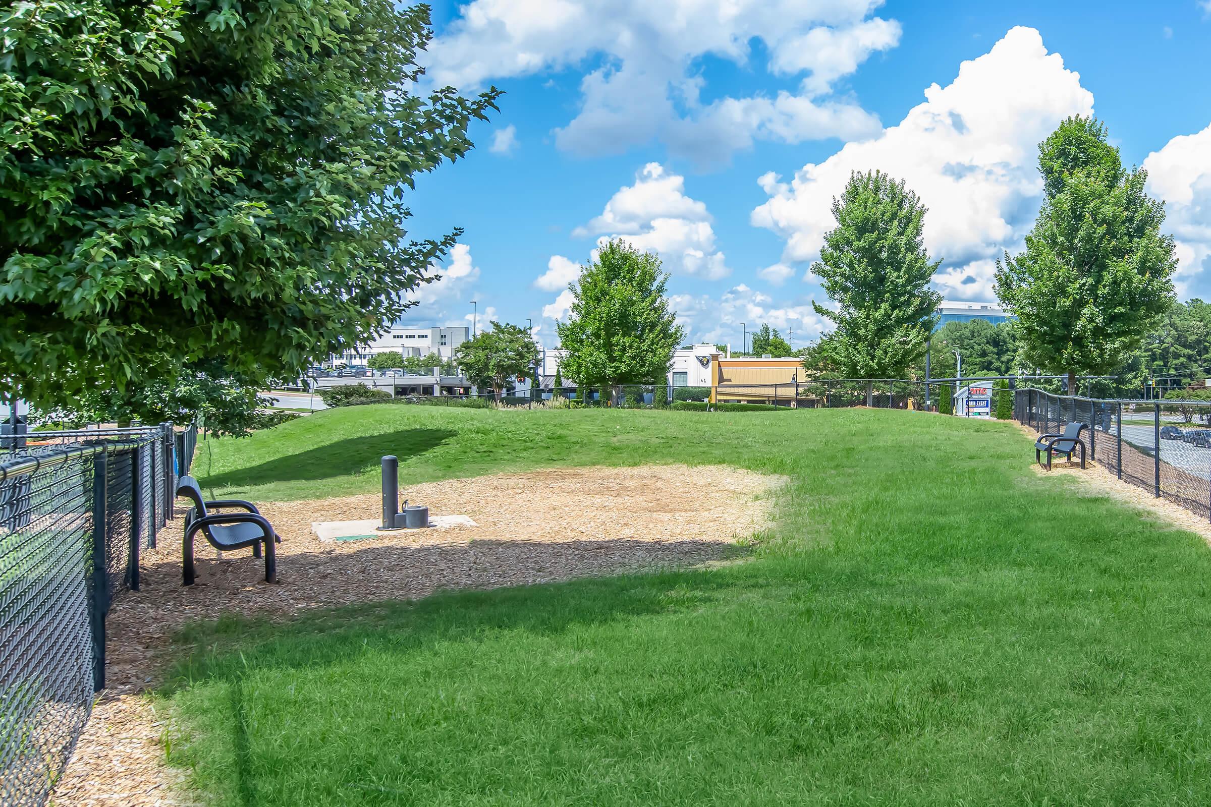 A small, grassy park area surrounded by a black fence. Two park benches are positioned on the side, and a water fountain for dogs is visible in the center. The sky is bright with fluffy clouds, and several trees provide shade in the area.