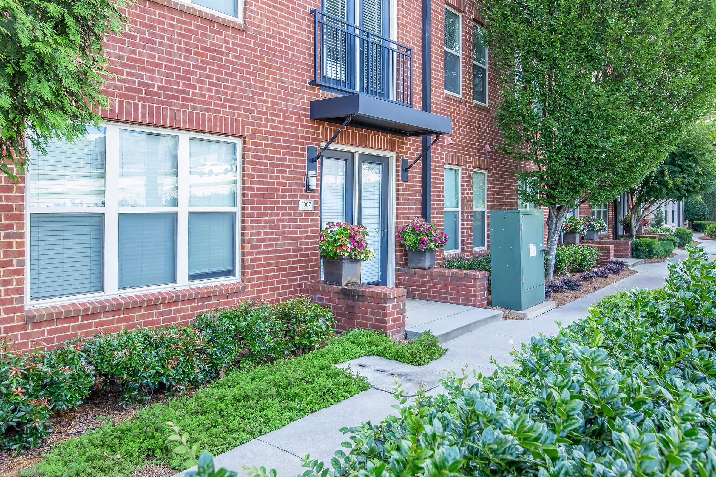 Exterior view of a modern brick apartment building with large windows. Colorful flower boxes hang from the balcony. The entrance features a small awning, and the surrounding landscape includes neatly trimmed bushes and trees. A sidewalk leads up to the entrance, and a utility box is visible nearby.