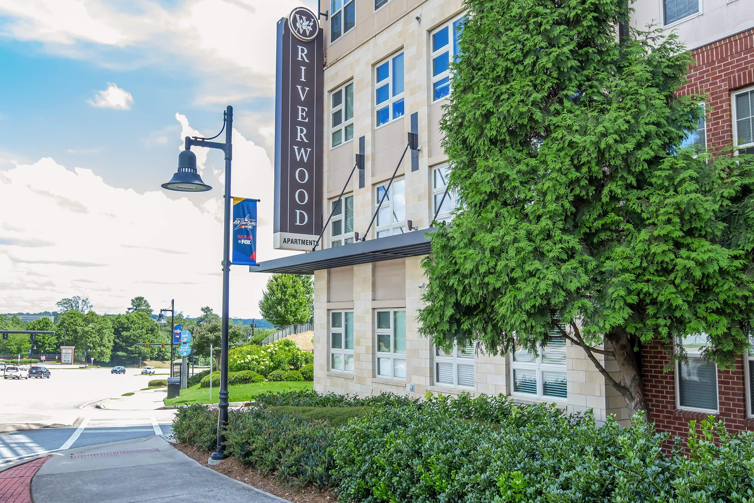 A view of the Riverwood Apartments building, featuring a prominent sign and lush greenery in the foreground. The architecture is modern, with large windows and a well-maintained landscape. Streetlights line the walkway, and a clear sky adds to the inviting atmosphere.