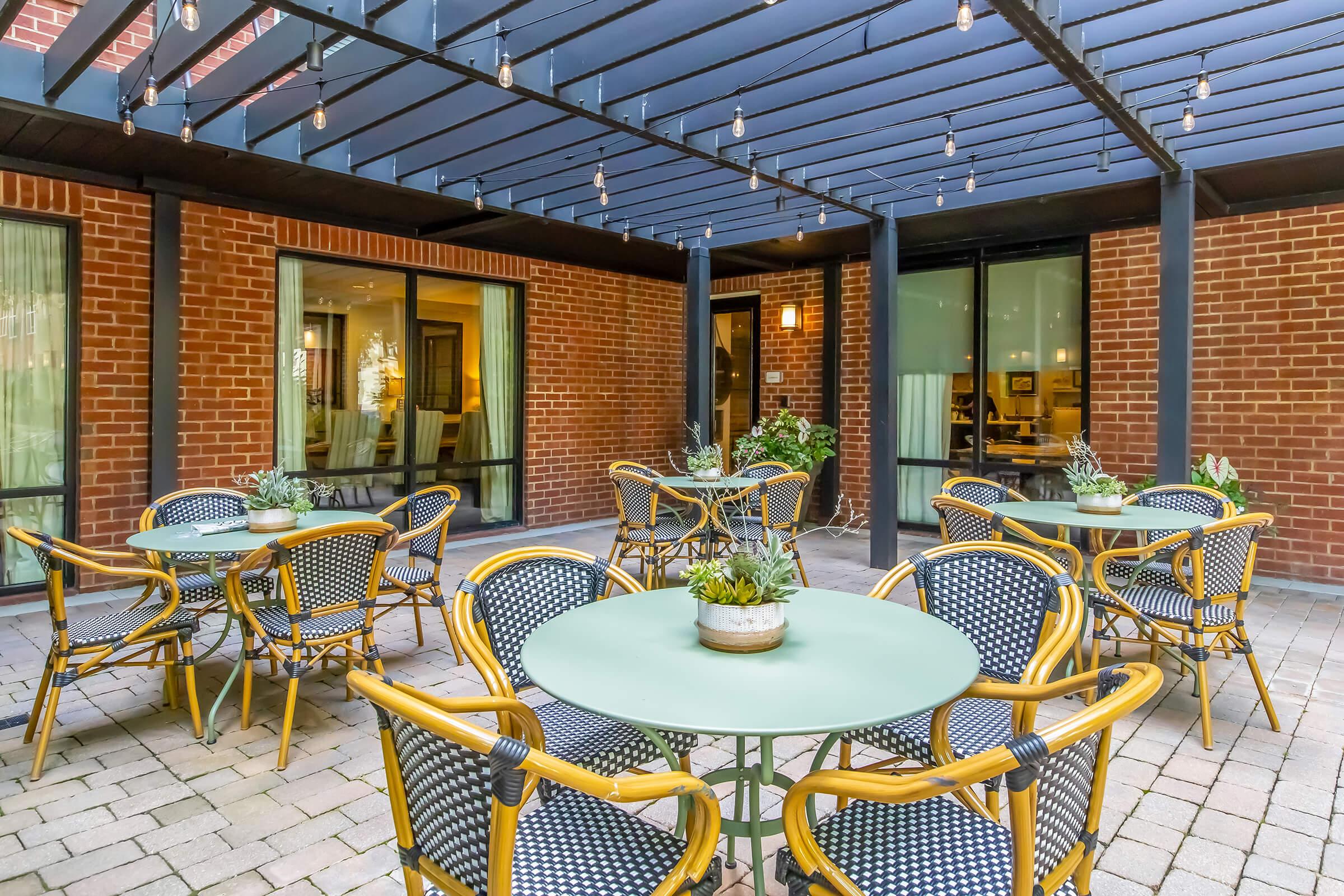 A cozy outdoor seating area featuring several round tables with green tops and rattan-style chairs. The space is shaded by a dark overhead structure, with string lights adding ambiance. Surrounding the tables are potted plants, set against a backdrop of brick walls and large windows.