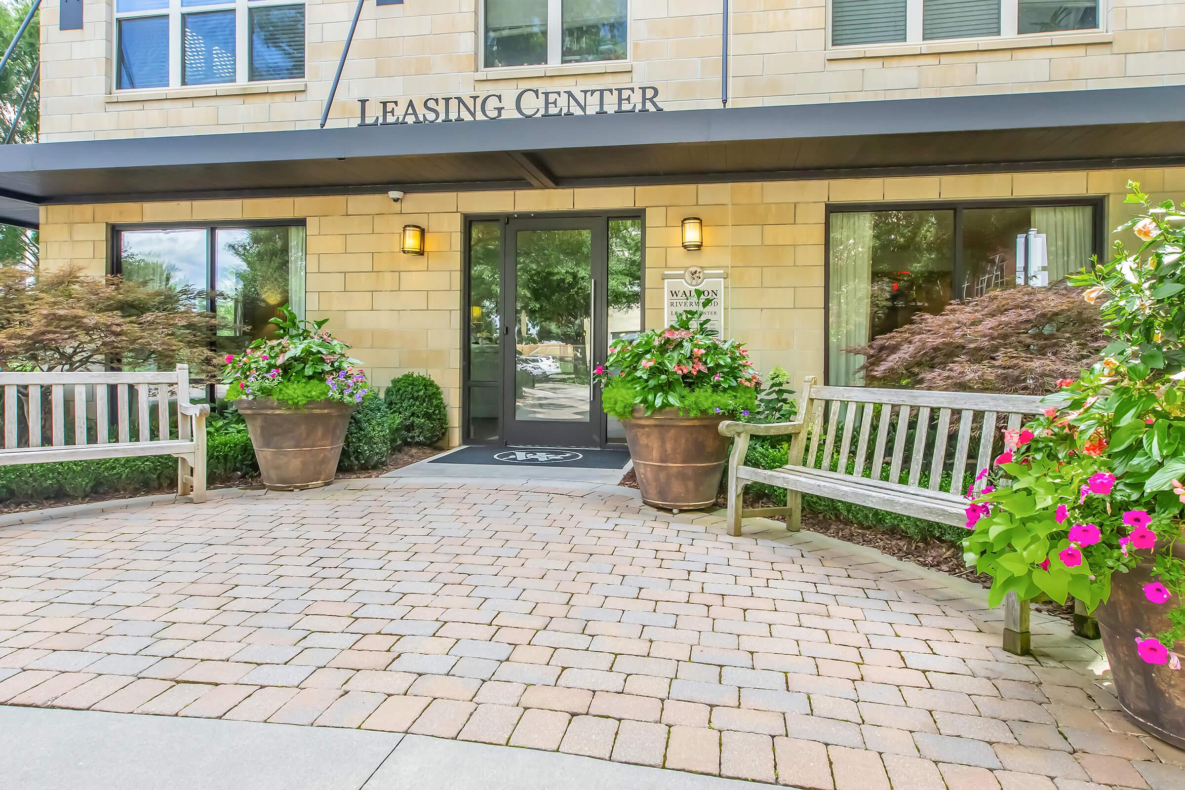 Exterior view of a leasing center featuring a stone facade, large windows, and a glass entrance door. Two wooden benches are placed on either side of the walkway, adorned with colorful flower pots. The area is landscaped with shrubs and flowers, creating an inviting atmosphere.