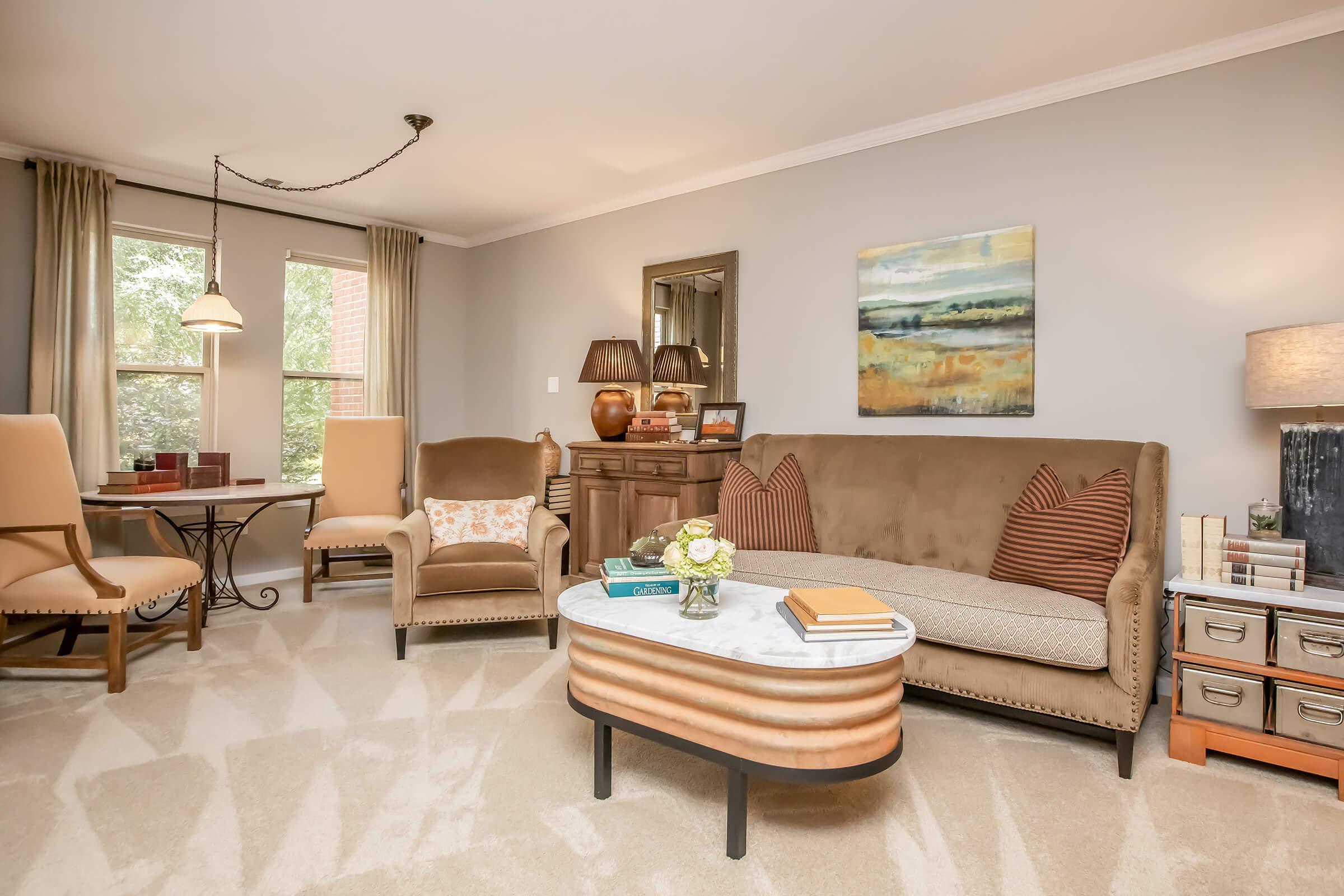A cozy living room featuring a brown sofa with patterned cushions, an accented armchair, and a round coffee table. There's a side table with decorative items, a landscape painting on the wall, and natural light coming through large windows, creating a warm and inviting atmosphere.