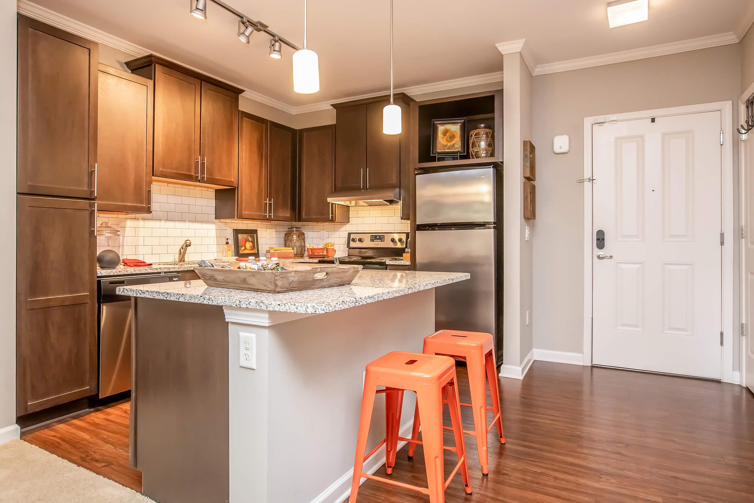 Modern kitchen featuring dark wood cabinets, stainless steel appliances, and a stylish island with three orange stools. The space is well-lit with overhead lights, has a contemporary design, and includes a door leading outside. The countertops are made of granite, and there is an inviting atmosphere.
