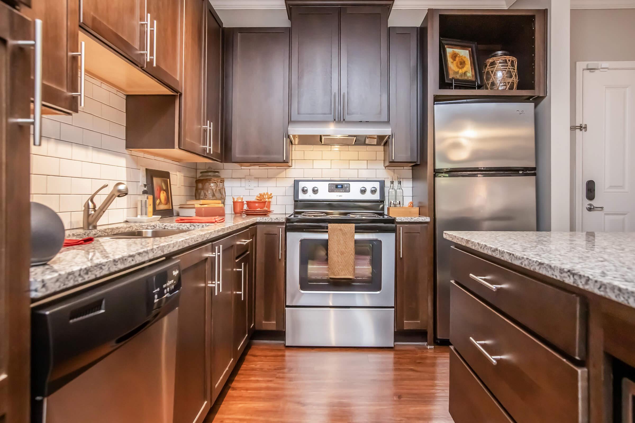 Modern kitchen featuring dark wooden cabinets, a stainless steel refrigerator, and an oven range. The countertops are made of granite, complemented by a backsplash of white tiles. The island has drawers and space for seating, while the overall design emphasizes a contemporary, inviting atmosphere.