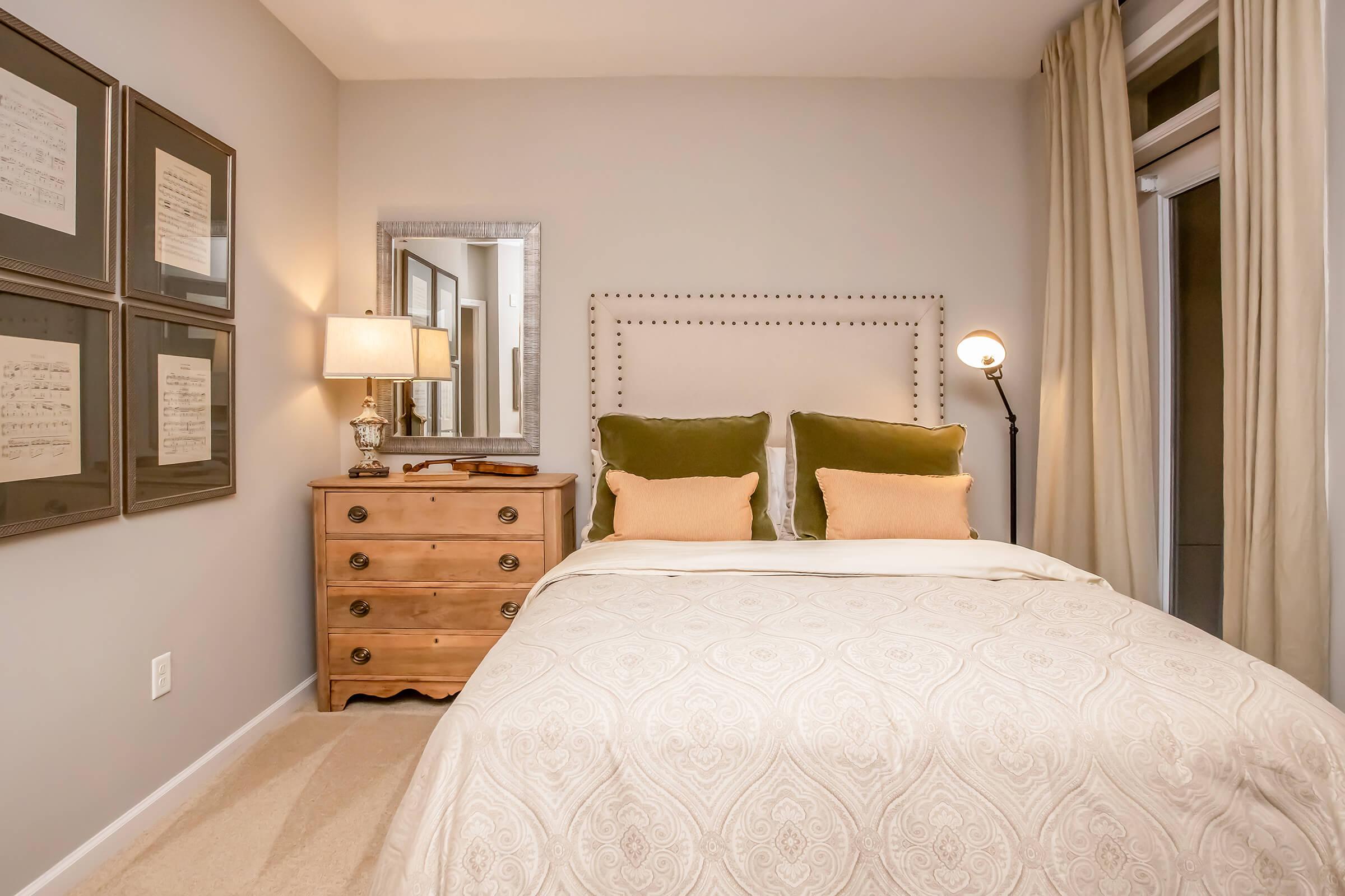 Cozy bedroom featuring a neatly made bed with a patterned white and beige comforter. The headboard has green velvet pillows. A wooden dresser is positioned next to the bed, with framed wall art above it. A table lamp and floor lamp provide warm lighting, and beige curtains frame the window.