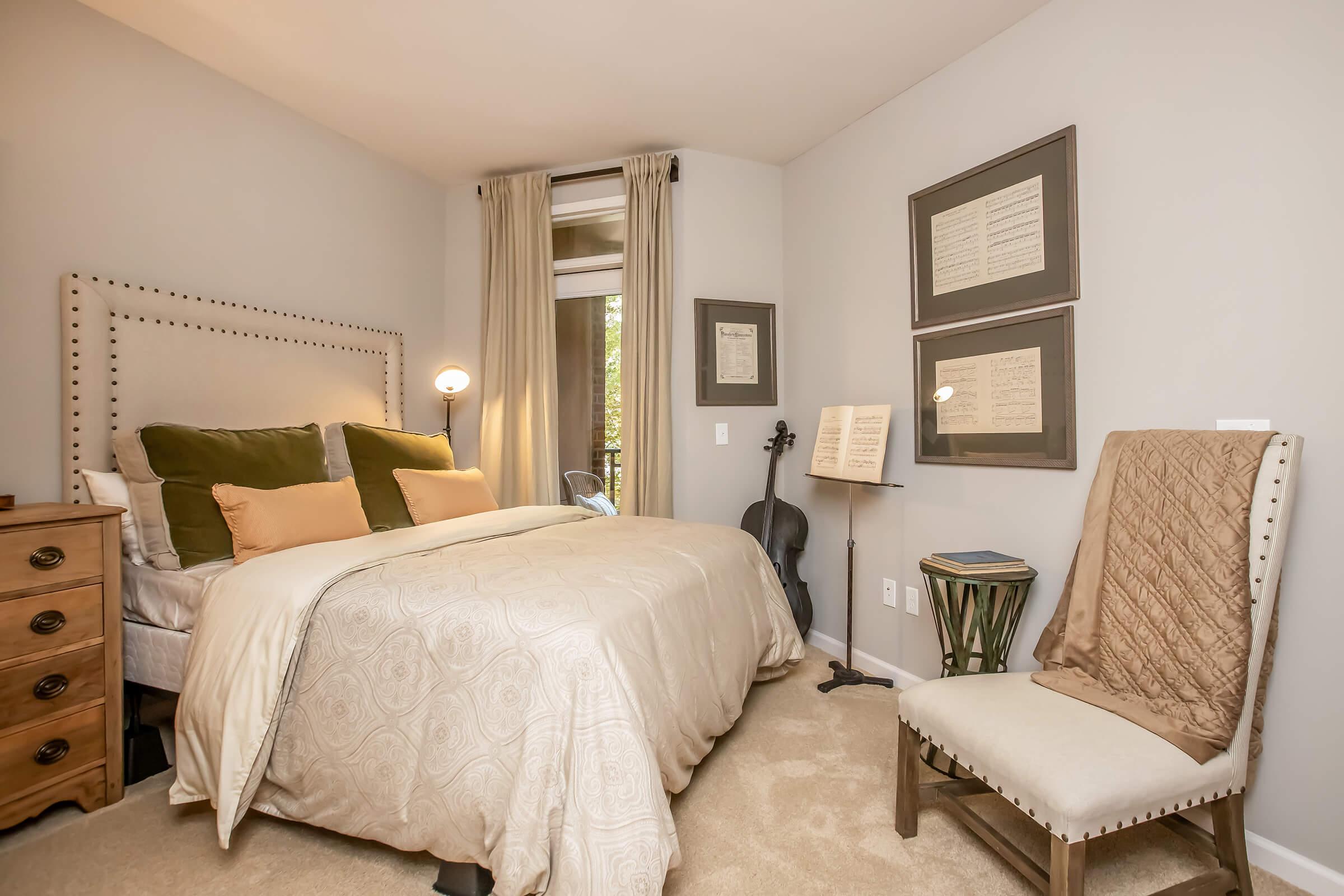 A cozy bedroom featuring a bed with decorative pillows, a bedside table, a music stand with sheets, and a chair. Soft lighting from a lamp and large windows bring warmth, while the walls display framed artwork. The carpeted floor adds to the inviting atmosphere.
