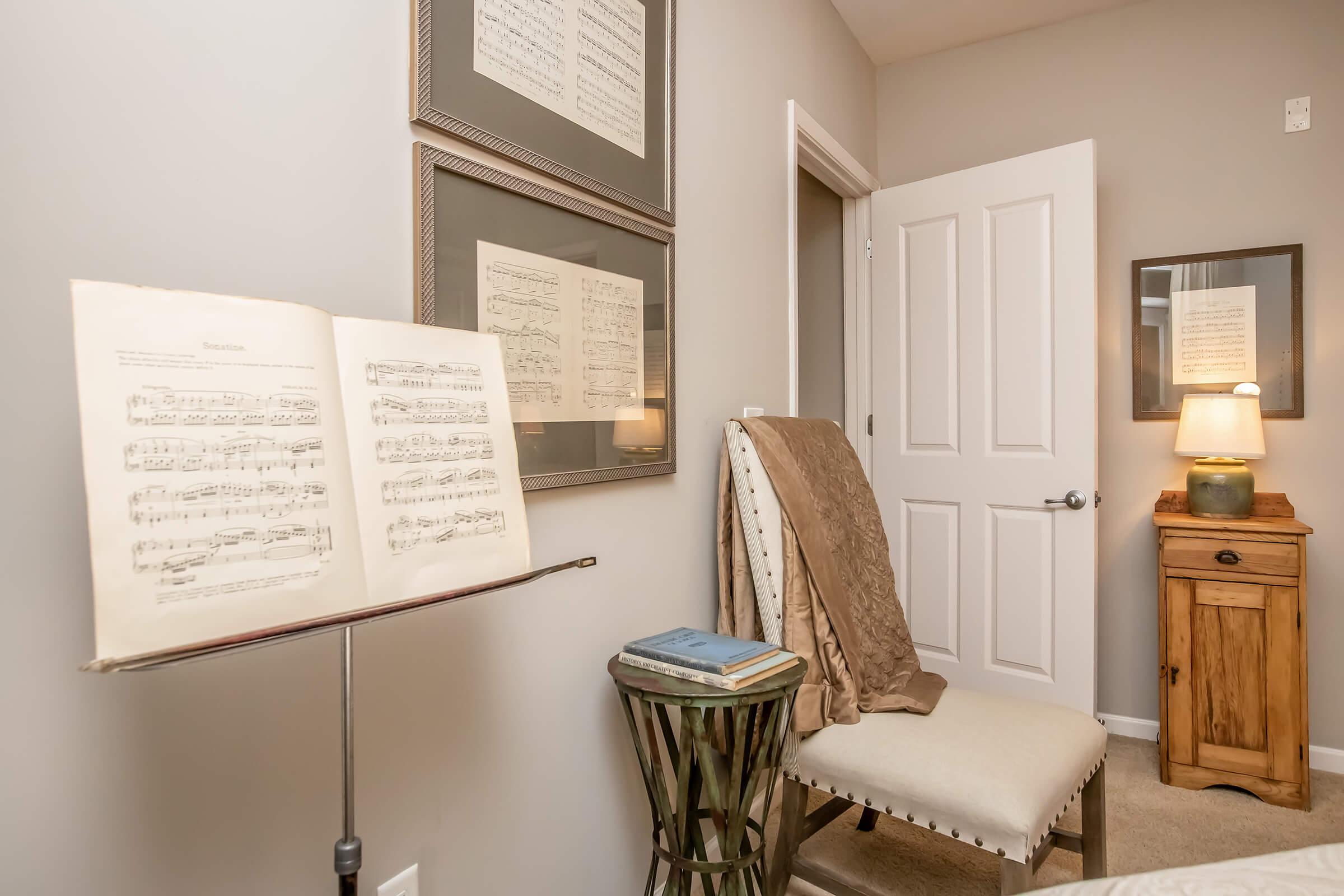 A cozy room featuring a music stand with sheet music, a chair draped in a blanket, and a wooden nightstand with a small lamp. The walls are adorned with framed sheet music, and a door leads to another space, enhancing the inviting atmosphere.