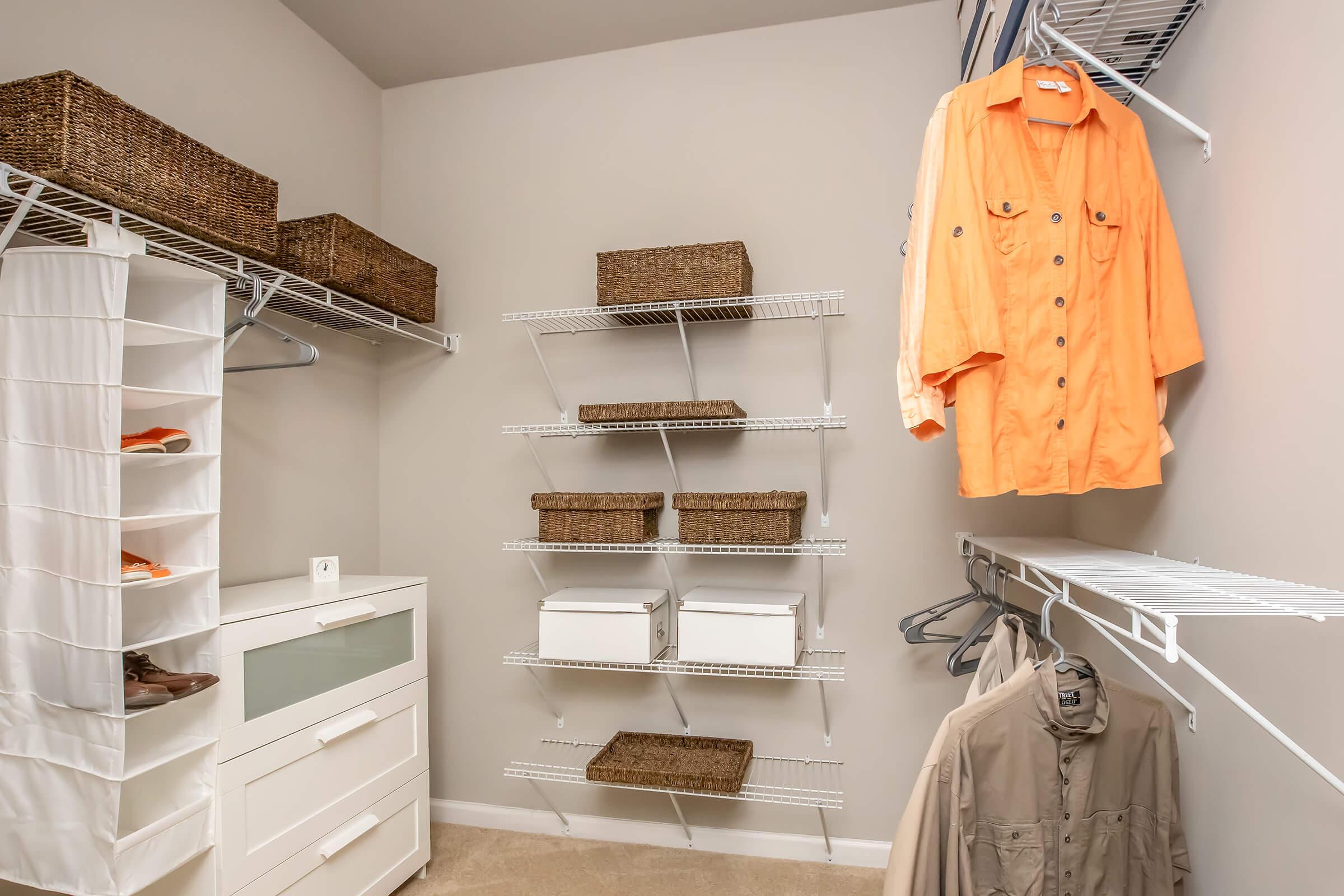 A spacious closet featuring white wire shelving, several woven storage baskets, and hanging clothes including an orange shirt and beige jacket. A white cabinet with drawers is positioned at the bottom, and the walls are painted a light color, creating a clean and organized space.