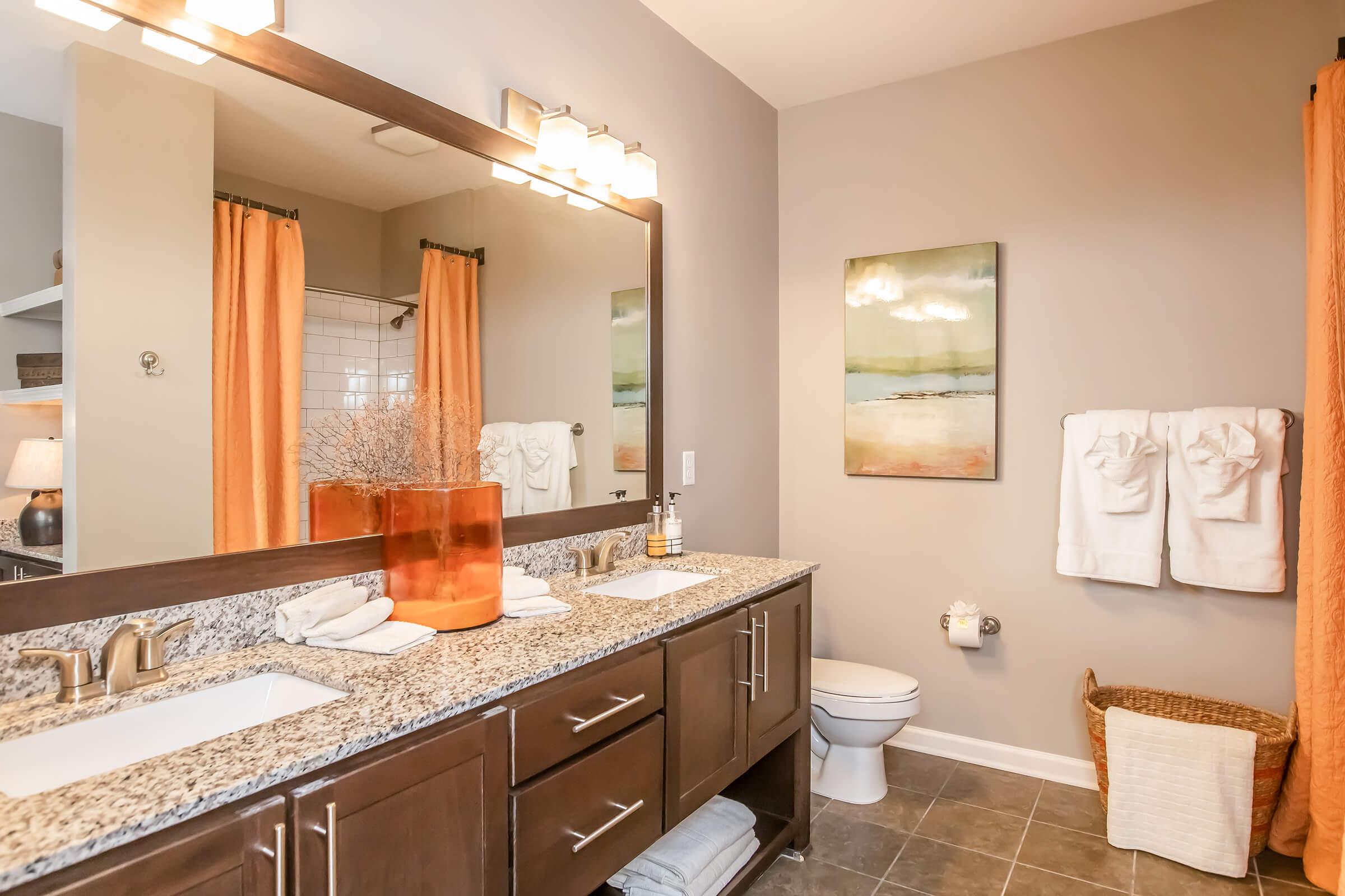 A modern bathroom featuring a double vanity with granite countertops, two sinks, and stylish faucets. The wall is painted in gray, adorned with a framed painting. Bright orange shower curtains complement the decor, and there are neatly hung towels. A clean toilet and a wicker basket add to the organized appearance.
