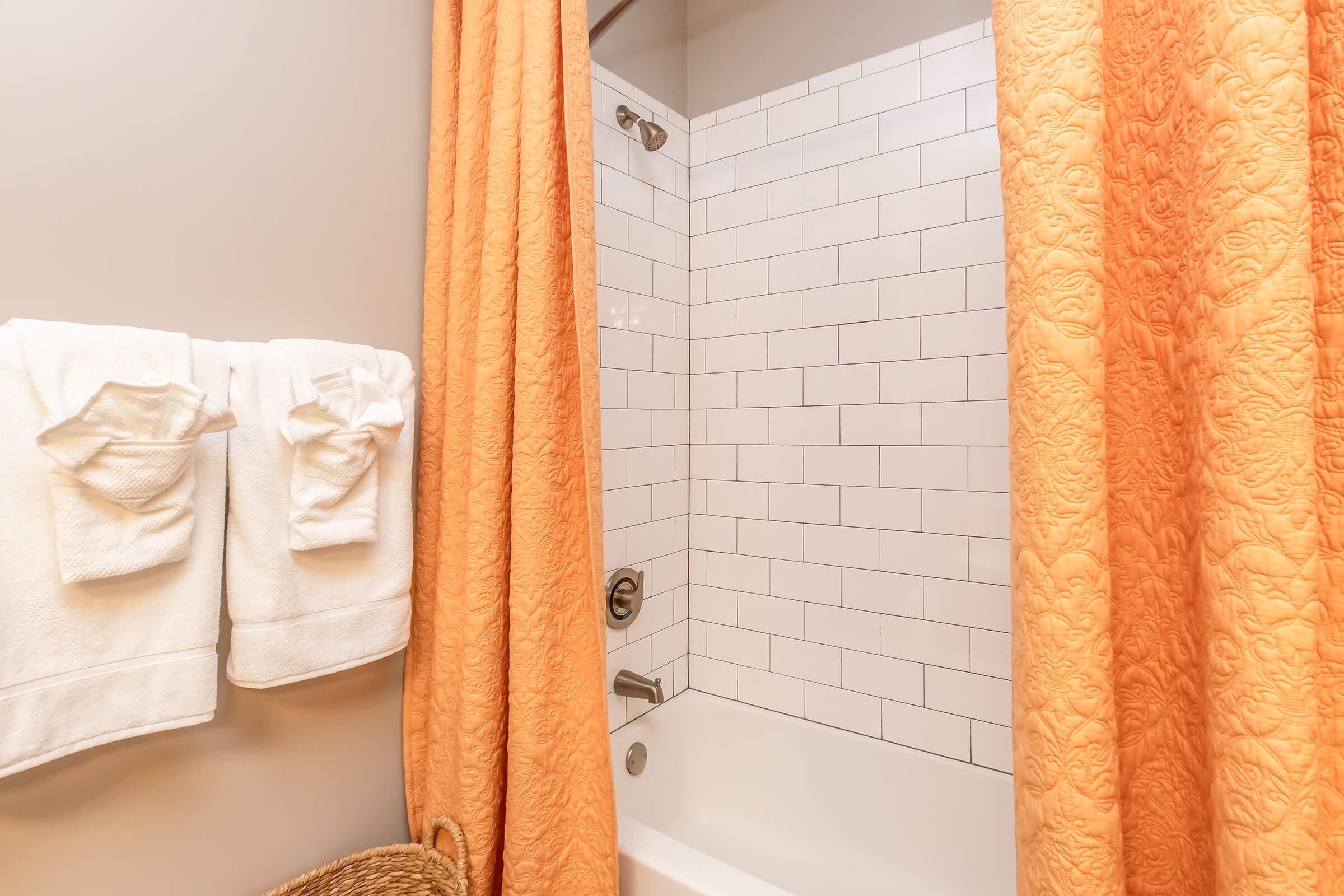 A bathroom with a white tiled shower and a bathtub. There is an orange patterned shower curtain partially covering the tub. Two white towels are neatly hung on a rack next to the shower. A woven basket is visible on the floor. The walls are a soft gray color.