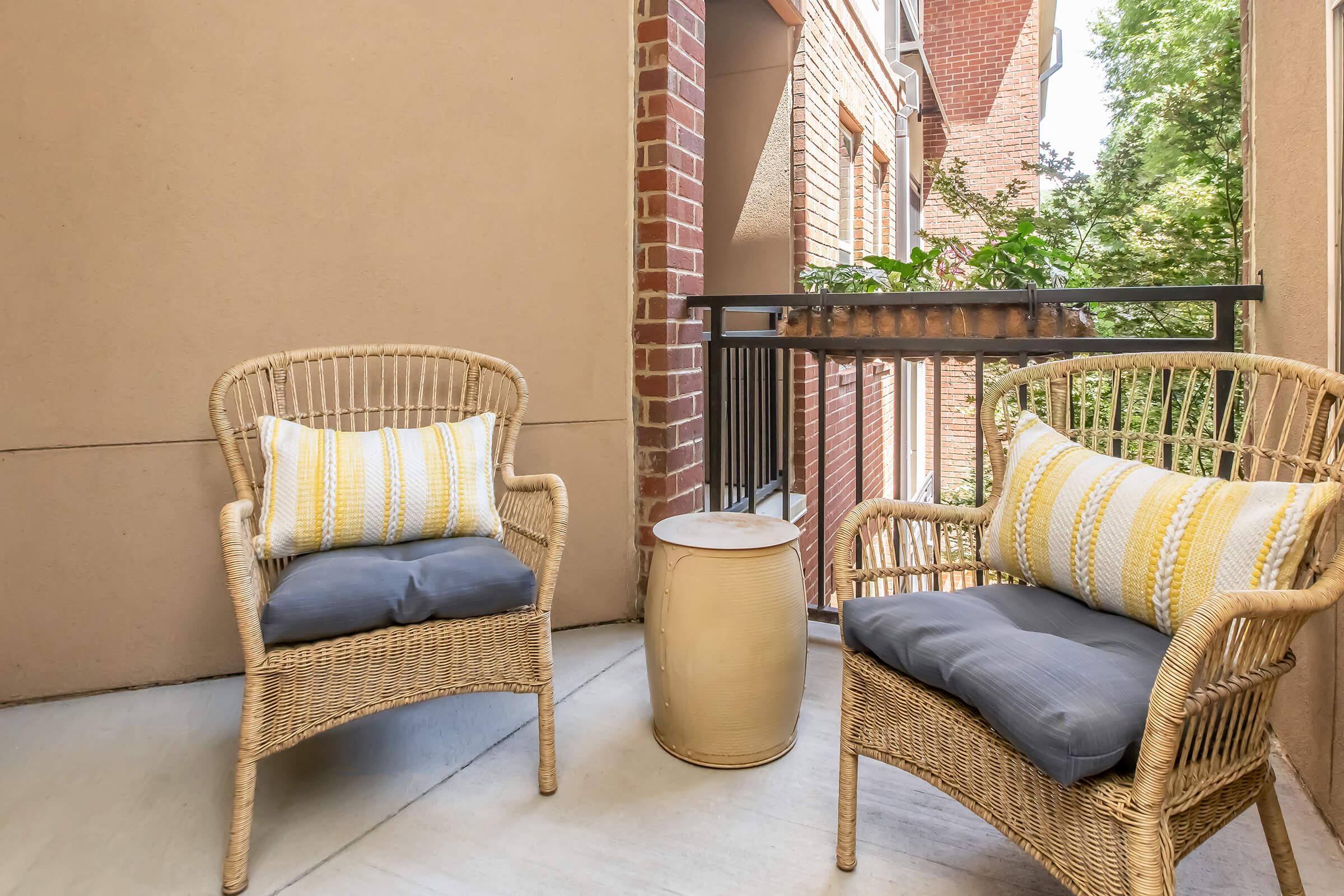 A cozy balcony setting featuring two wicker chairs with cushions, a small round table, and a railing adorned with green plants. The wall is a light beige color, complementing the brickwork of the building in the background. The atmosphere is inviting and perfect for relaxing outdoors.