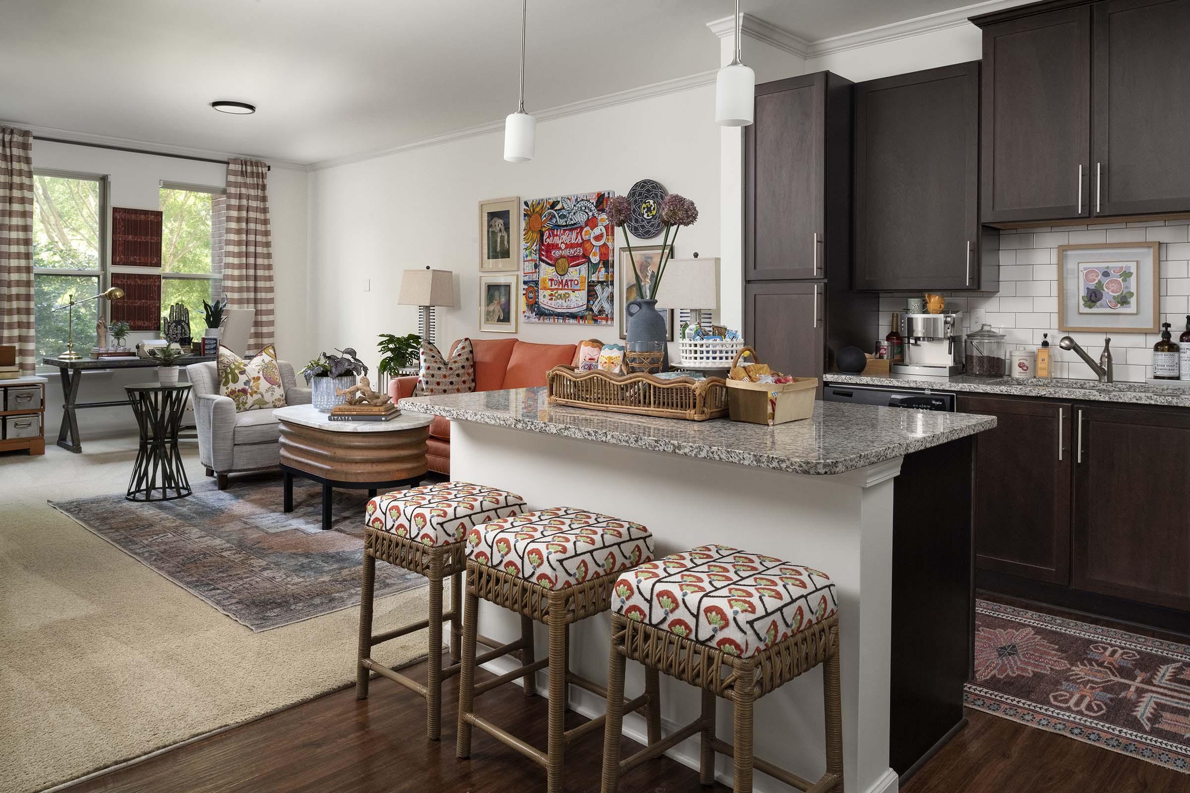 A modern living space featuring a cozy kitchen and dining area. The kitchen has dark wood cabinets, a granite countertop, and bar stools with a patterned fabric. An orange sofa is placed in the living area, which includes a coffee table and a colorful rug. Large windows provide natural light, enhancing the inviting atmosphere.
