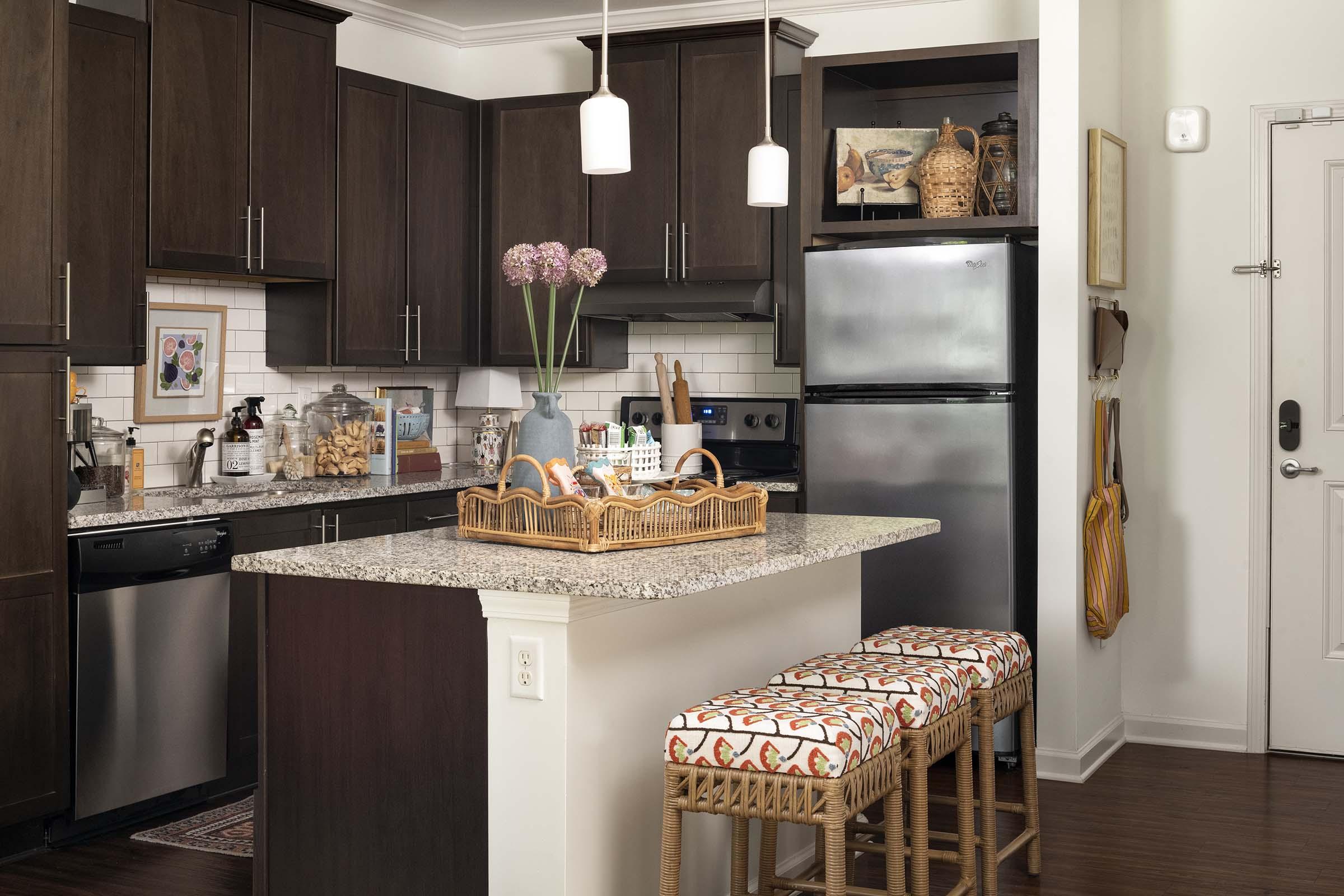 Modern kitchen featuring dark wood cabinetry, a granite countertop island with decorative baskets, and stylish bar stools. There is a stainless steel refrigerator and an oven, with a small potted plant and colorful flowers on the island. Light fixtures hang above, and a door is visible in the background.