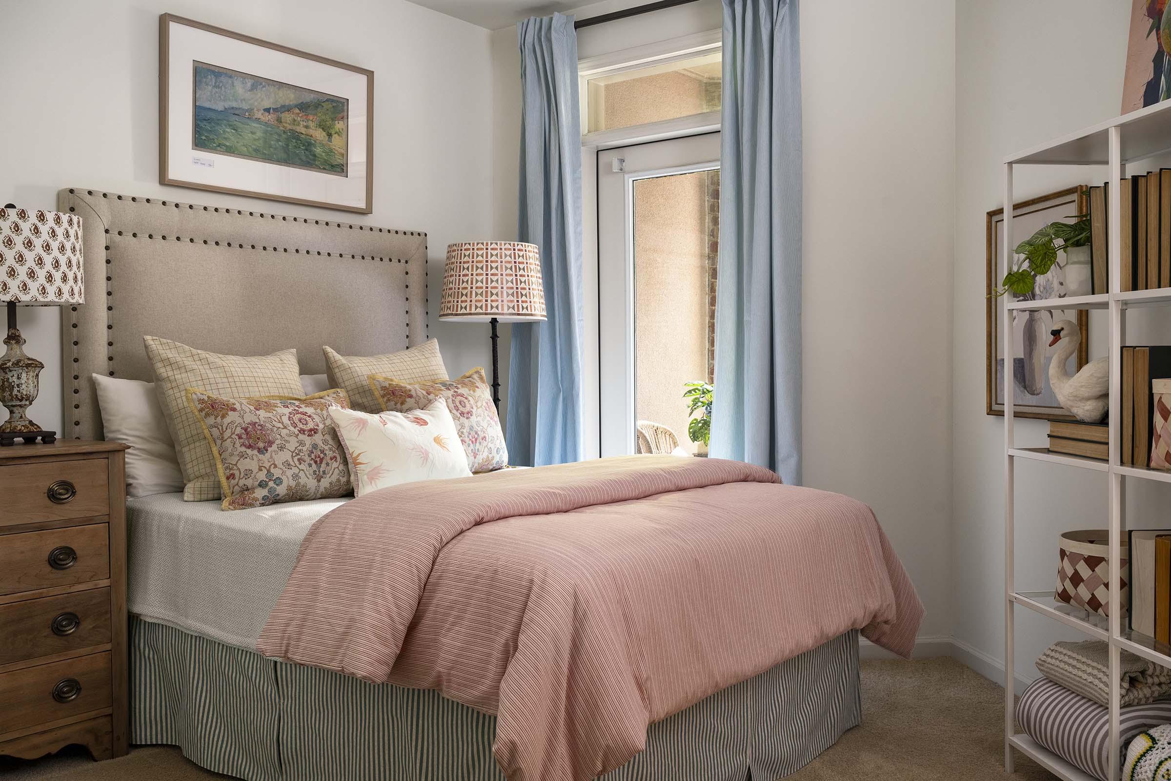 A cozy, well-decorated bedroom featuring a plush bed with decorative pillows and a soft pink blanket. There is a wooden nightstand with a lamp, a large window with blue curtains, and a bookshelf filled with framed art and decorative items. The overall aesthetic is warm and inviting.