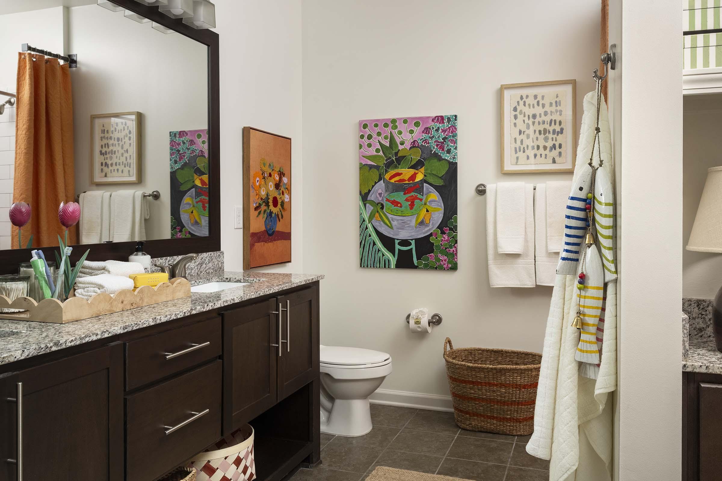 A modern bathroom featuring a dark wooden vanity with a granite countertop, decorated with neatly arranged toiletries and towels. There are two colorful artworks on the walls, one depicting flowers and the other a vibrant still life. A basket and a striped towel hang nearby, creating a cozy atmosphere.