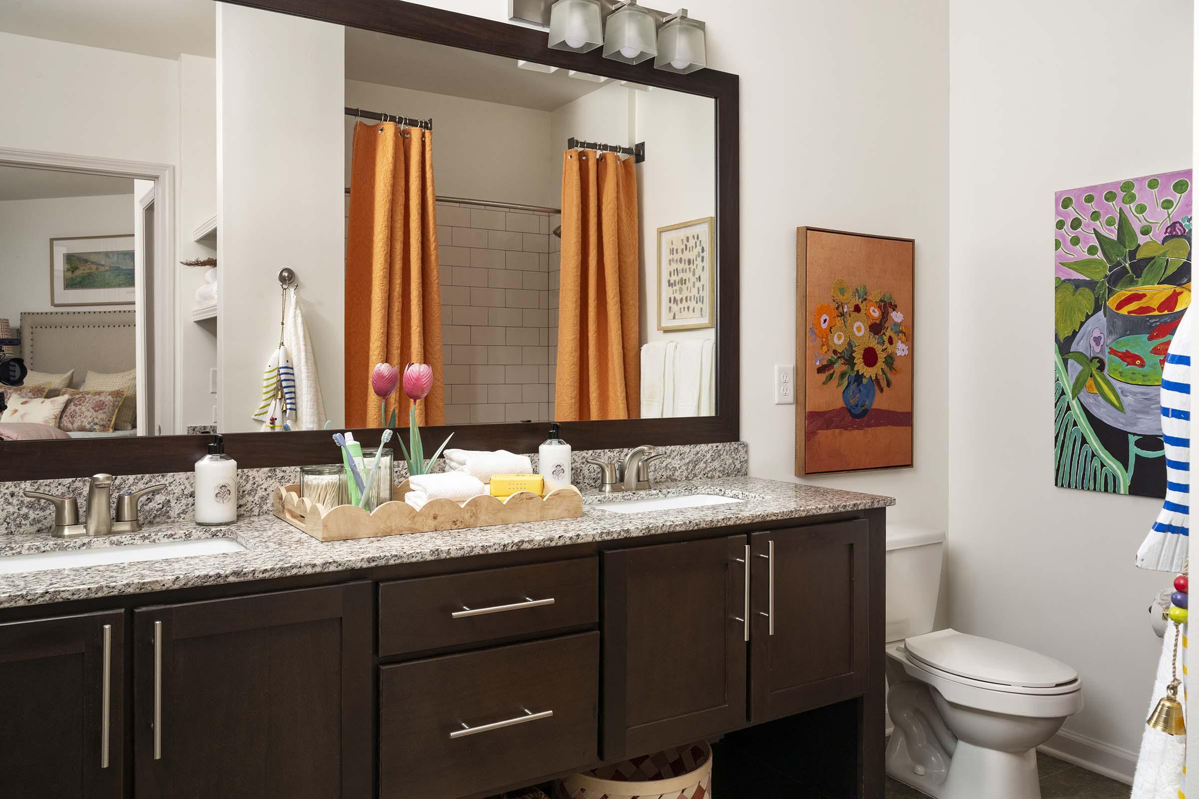 A modern bathroom featuring a double vanity with a granite countertop, two sinks, and decorative containers. The decor includes orange shower curtains, a framed floral painting, and a colorful towel rack. A glimpse of a bedroom is visible in the background. Bright and inviting atmosphere.