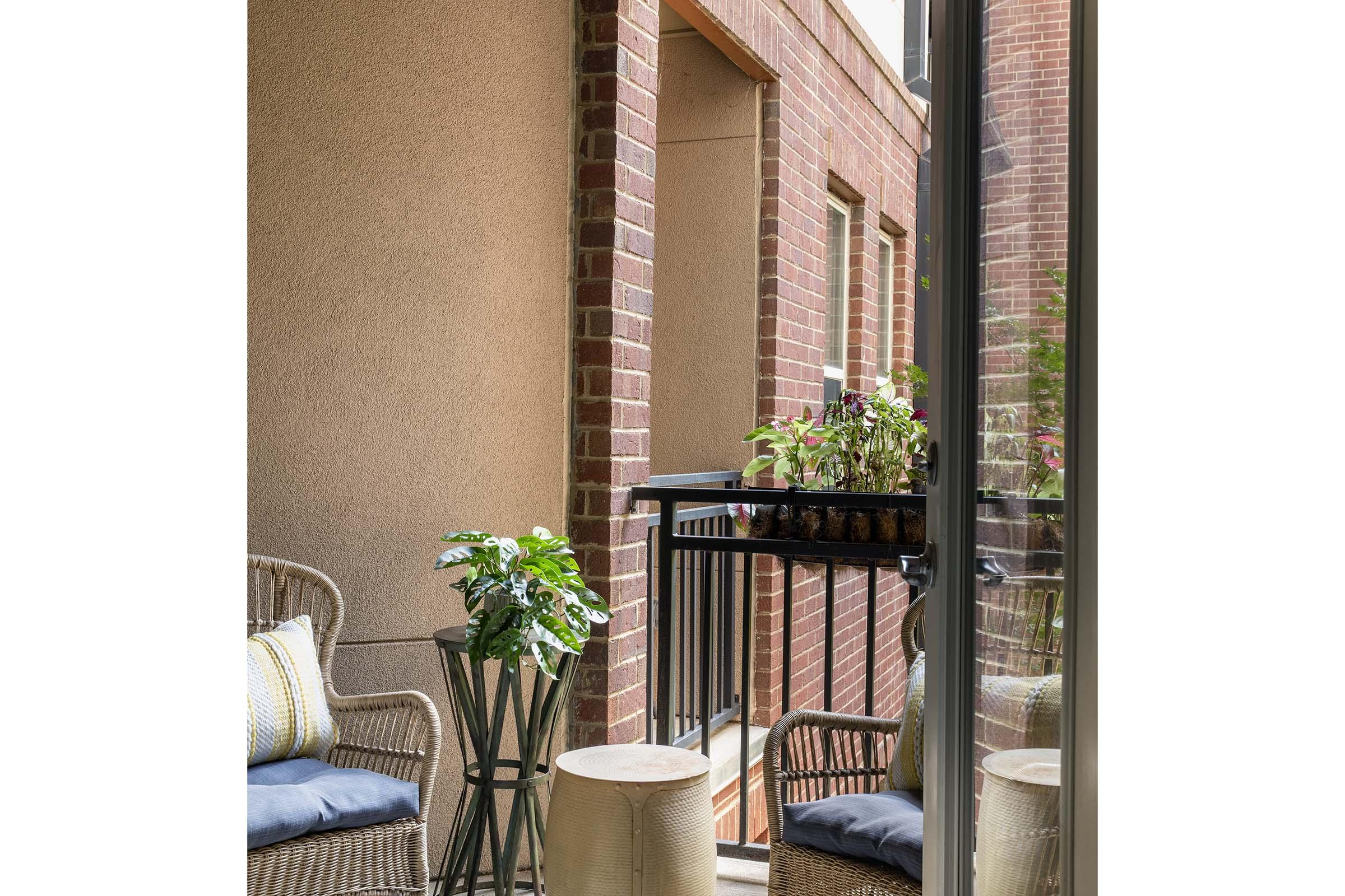 A cozy balcony featuring two wicker chairs with cushions, a small round stool, and a decorative plant on a stand. There's a railing with potted plants, surrounded by brick walls, creating a warm and inviting outdoor space. The view reflects a sense of relaxation and comfort.