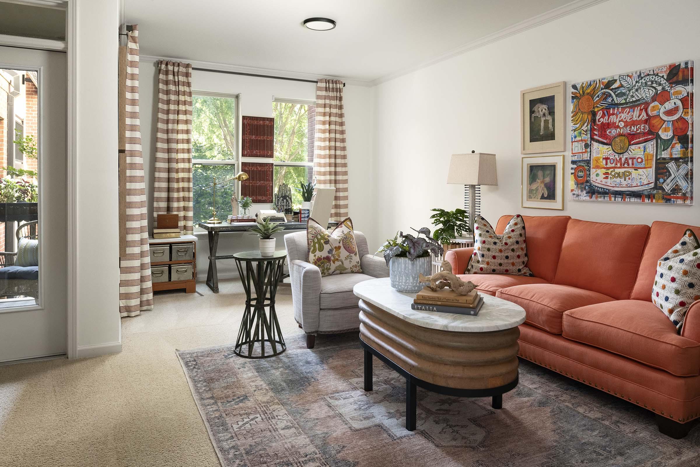 A cozy living room featuring an orange sofa, a light-colored armchair with decorative pillows, and a round coffee table. There are plants, framed artwork on the walls, and large windows with striped curtains, allowing natural light to fill the space. The decor is inviting and colorful, creating a warm atmosphere.