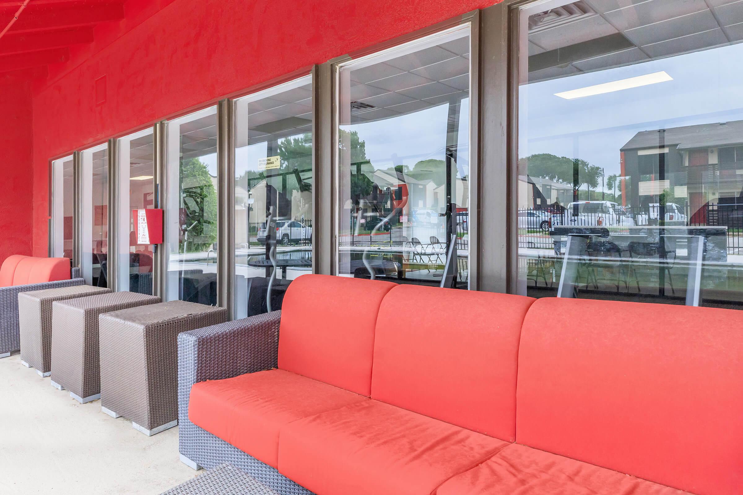 A red sofa in front of large windows, reflecting the outdoor area. The setting has modern furniture and a clean, contemporary design, suggesting a vibrant and inviting atmosphere. A pool and other buildings are visible through the windows in the background.