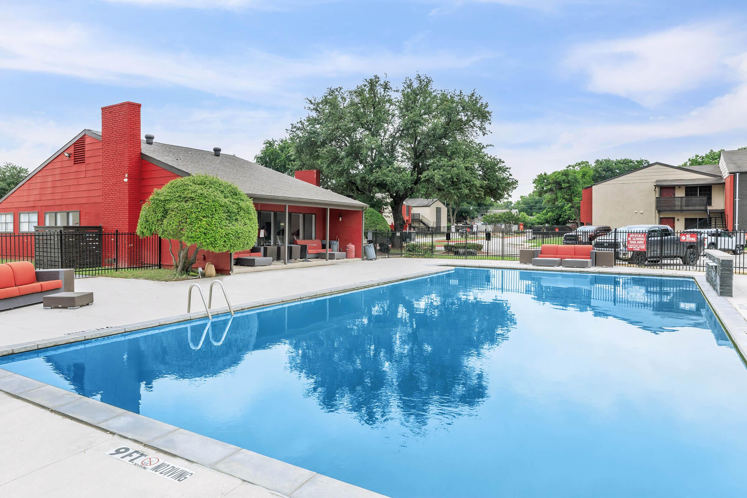 A clear swimming pool surrounded by a patio with lounge chairs and greenery. In the background, there is a red-brick building with large windows and seating areas, along with additional buildings visible. A sunny sky completes the scene, conveying a relaxing outdoor atmosphere.