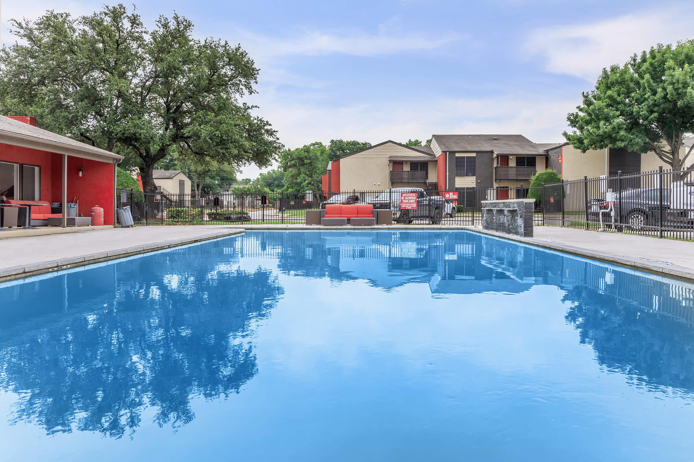 A clear blue swimming pool reflecting the sky and surrounding trees, with a fence in the background. Nearby, there are two apartment buildings visible, along with lounge seating near the pool. The scene conveys a peaceful outdoor space perfect for relaxation.