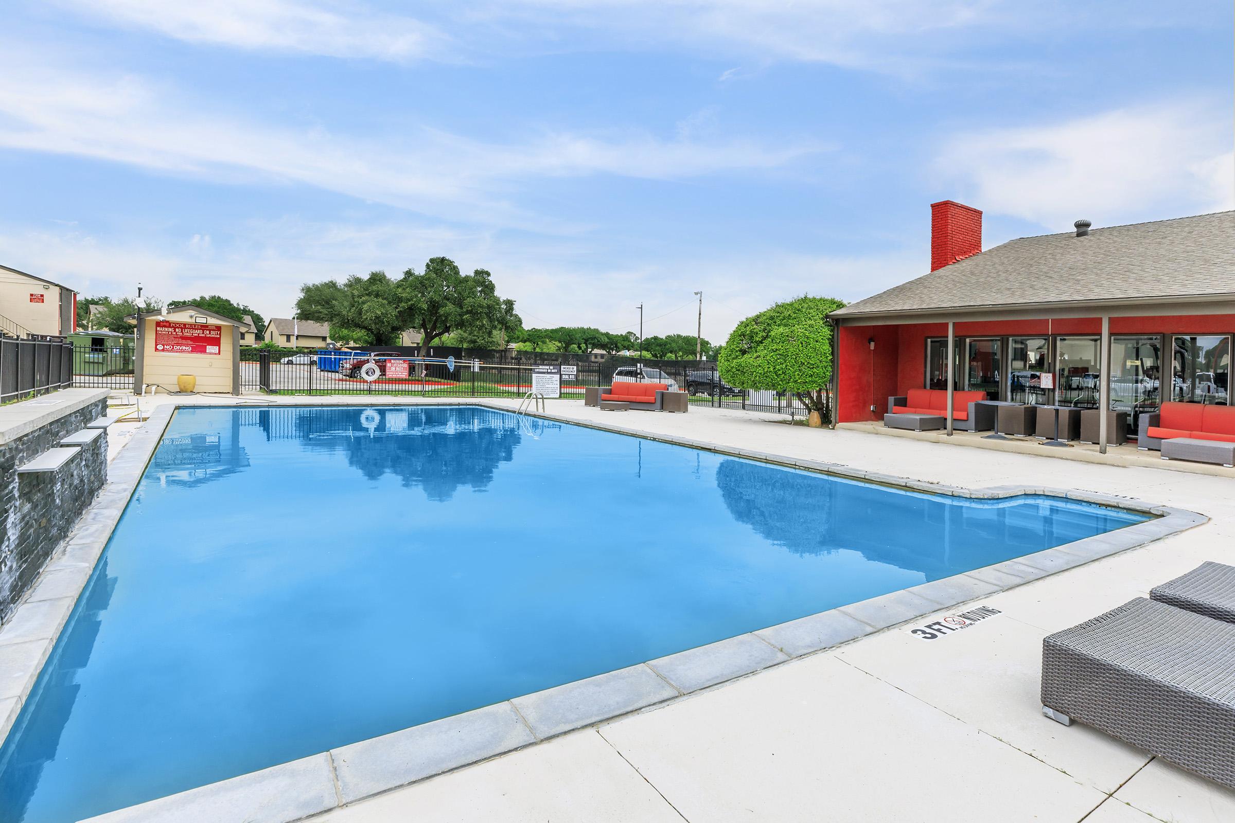 A clean, blue swimming pool surrounded by lounge chairs and shaded areas, with a modern building featuring large windows in the background. Nearby, there are green trees and an outdoor seating area, creating a relaxing atmosphere. The pool has clear water and a depth marker visible along the edge.