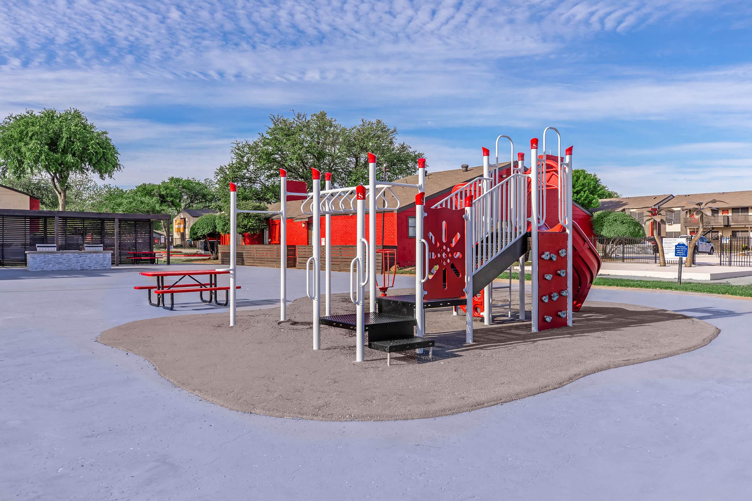 A colorful playground structure featuring a slide and climbing elements, surrounded by a smooth, gray play surface. Nearby, there are picnic tables under shaded trees, with a backdrop of residential buildings and a clear blue sky.