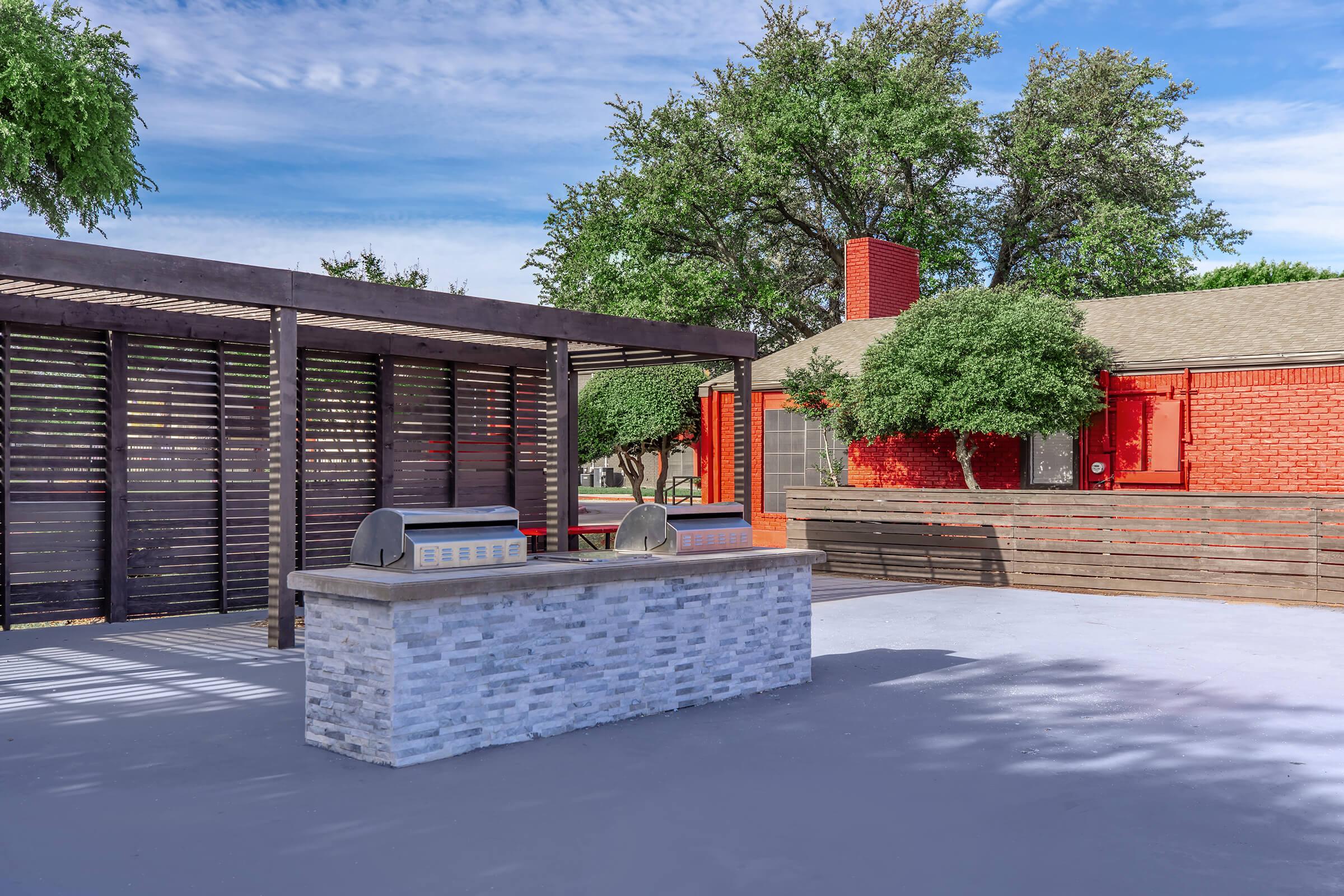 Outdoor patio area featuring a stone countertop with built-in grills, underneath a wooden pergola. Lush green trees and a red brick house are in the background, with a clear blue sky and scattered clouds above. The space is designed for outdoor cooking and entertaining.