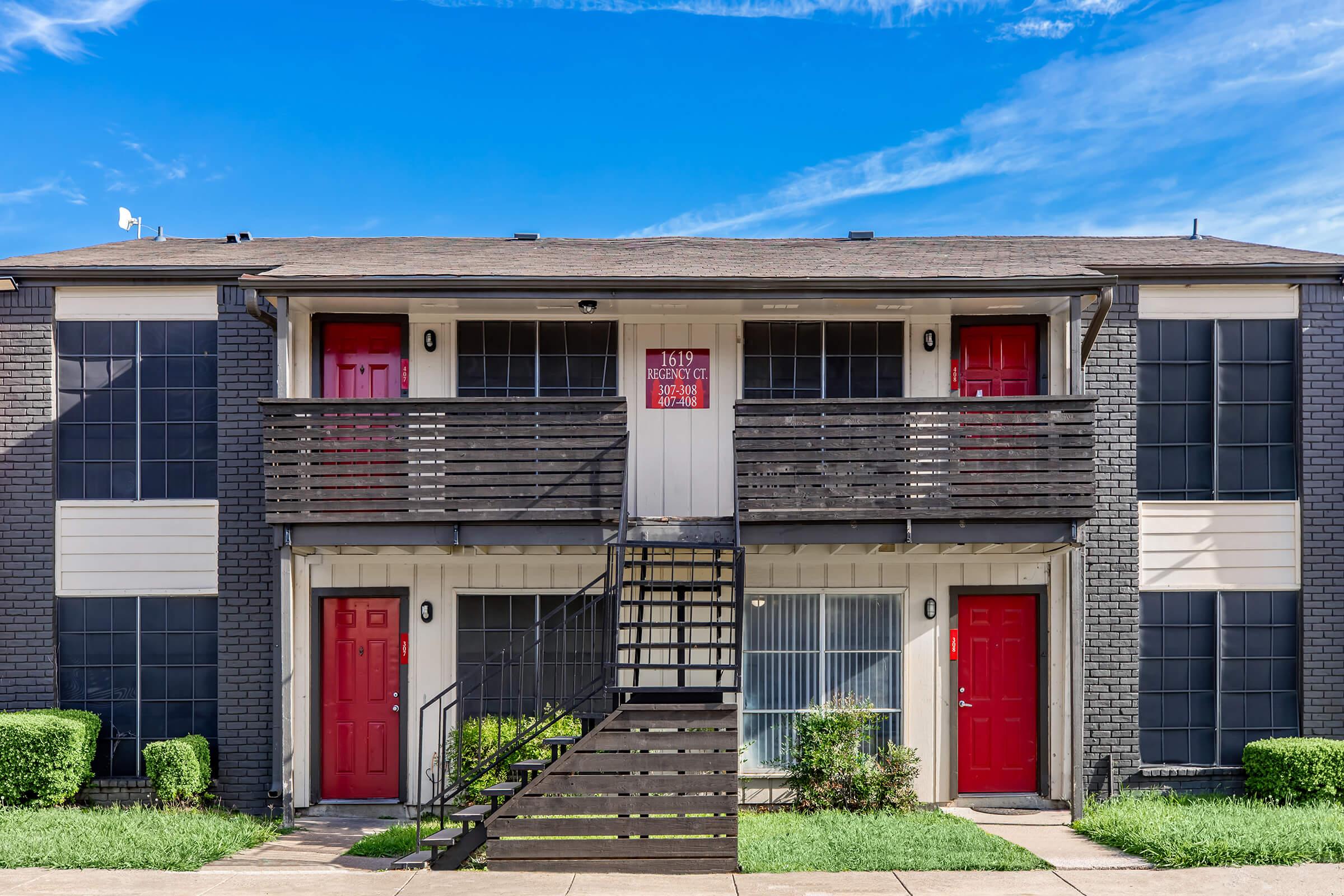 A two-story apartment building featuring a modern design, with two red doors on the ground and first floors. A wooden staircase leads to the upper unit. The building has large windows and is surrounded by well-maintained greenery under a bright blue sky.