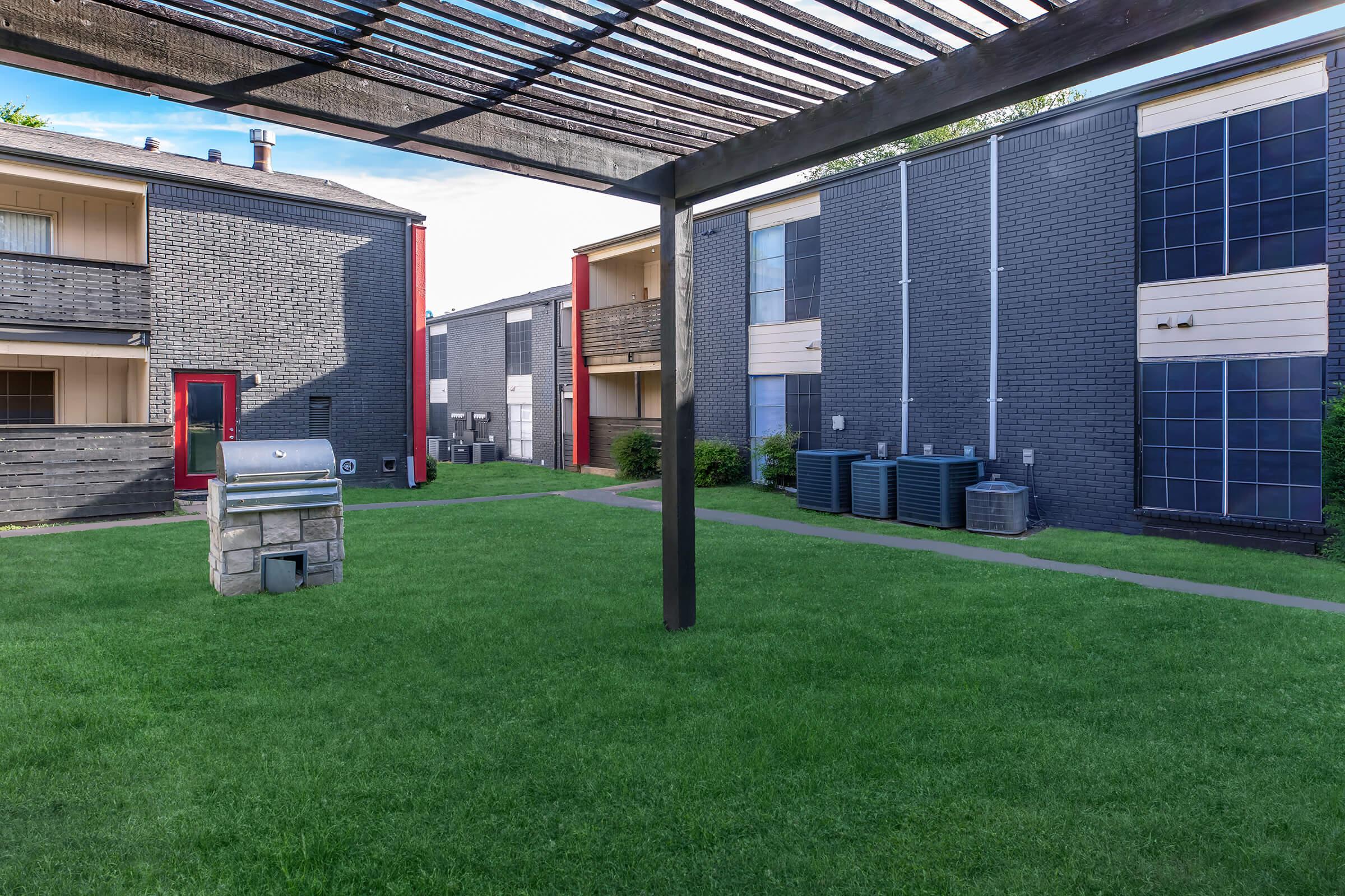 View of a grassy courtyard surrounded by apartment buildings. The foreground features a stone grill under a pergola, while several air conditioning units line the side of another building. The buildings are painted in shades of gray and feature red accents, with a clear blue sky overhead.