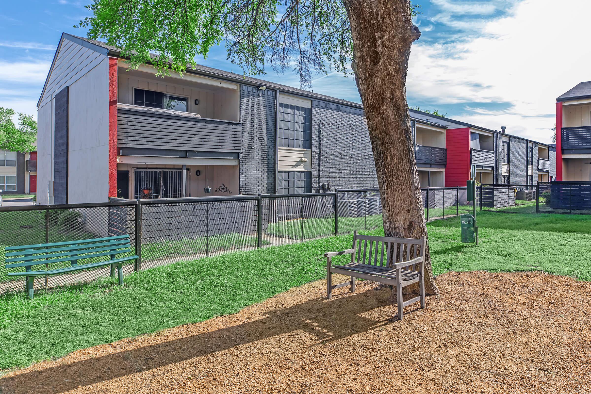 A view of a residential complex featuring two-story buildings with a mix of red and gray exteriors. There is a grassy area in the foreground with a wooden bench and a picnic table surrounded by gravel. A tree provides shade, and a fenced area is visible in the background. The sky is partly cloudy.