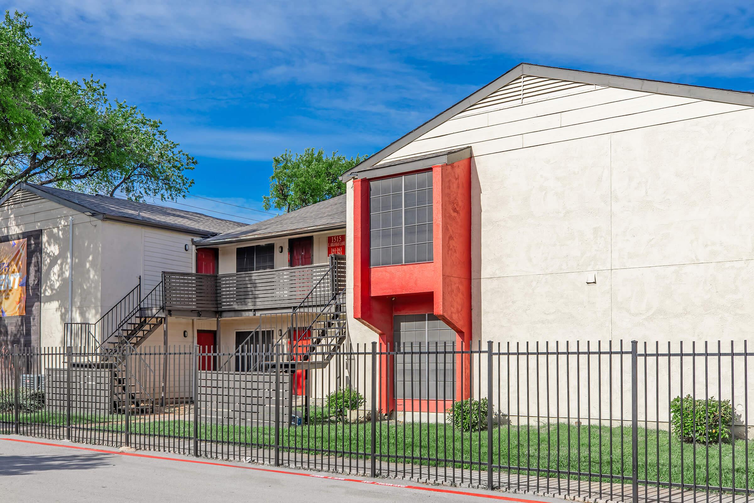 A two-story apartment building with a light-colored exterior and red-accented sections. The building features a staircase leading to the second floor, surrounded by a lawn. A black metal fence encloses the property, with a clear blue sky in the background.