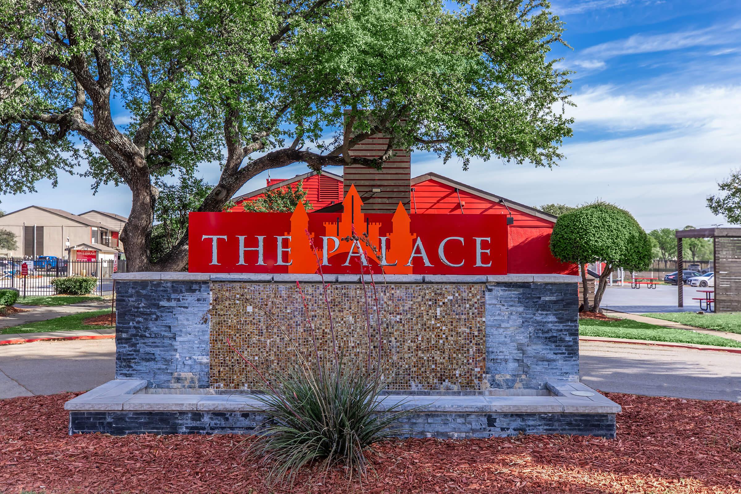 A decorative sign for "The Palace," featuring a modern design with red and gray elements. The sign is surrounded by lush green trees and landscaping, with a water feature in front. A blue sky enhances the vibrant setting, indicating a welcoming location.