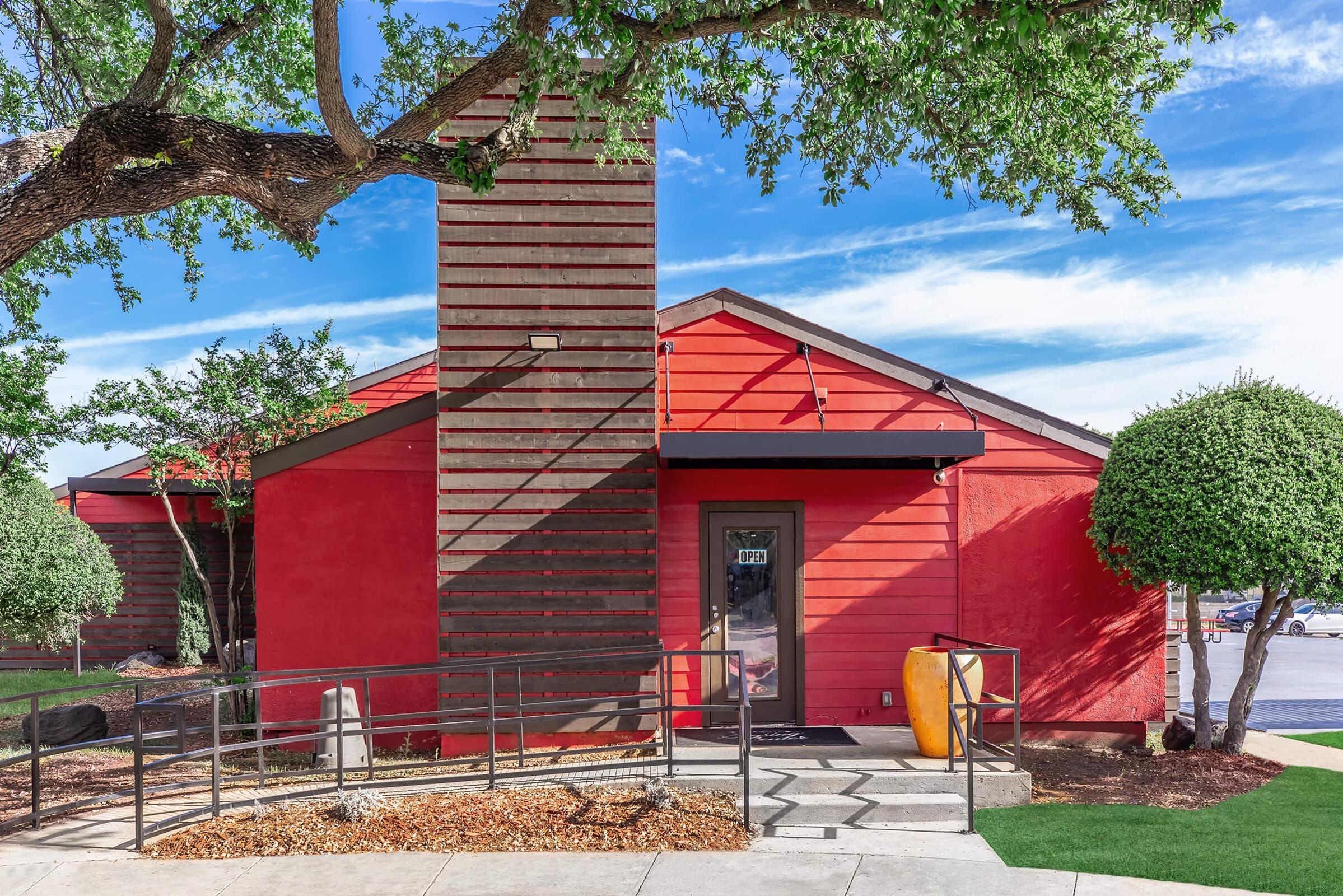 A modern red building with wooden accents, featuring a chimney, large windows, and a welcoming entrance. The area is landscaped with trees and trimmed bushes. A ramp leads to the entrance, making it accessible. The sky is bright with clouds, enhancing the vibrant atmosphere.