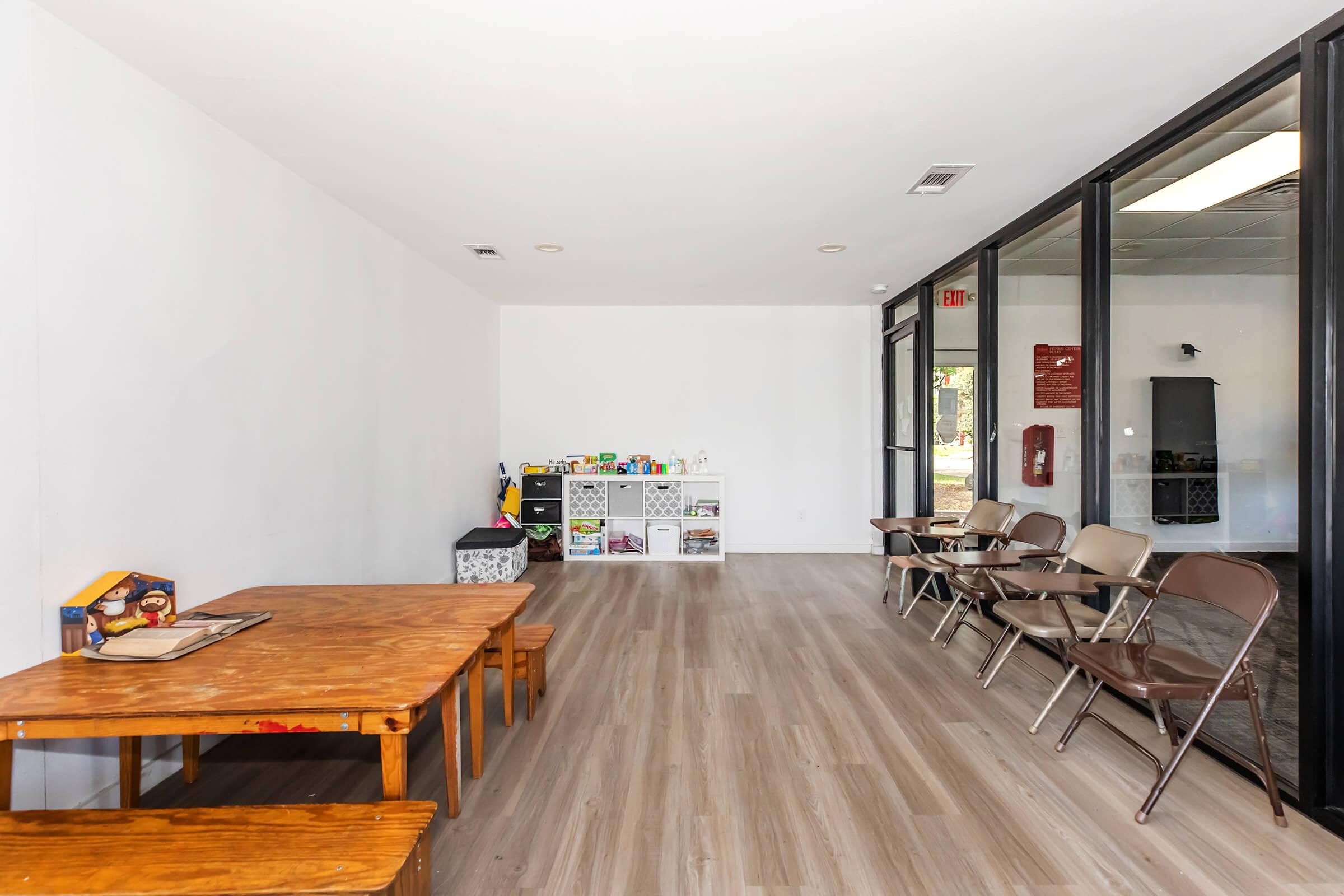 Interior view of a spacious room featuring two wooden tables and several metal folding chairs arranged in a line. The walls are painted white, and there is a small shelf filled with various items. Large windows allow natural light to illuminate the space, creating a bright and open atmosphere.